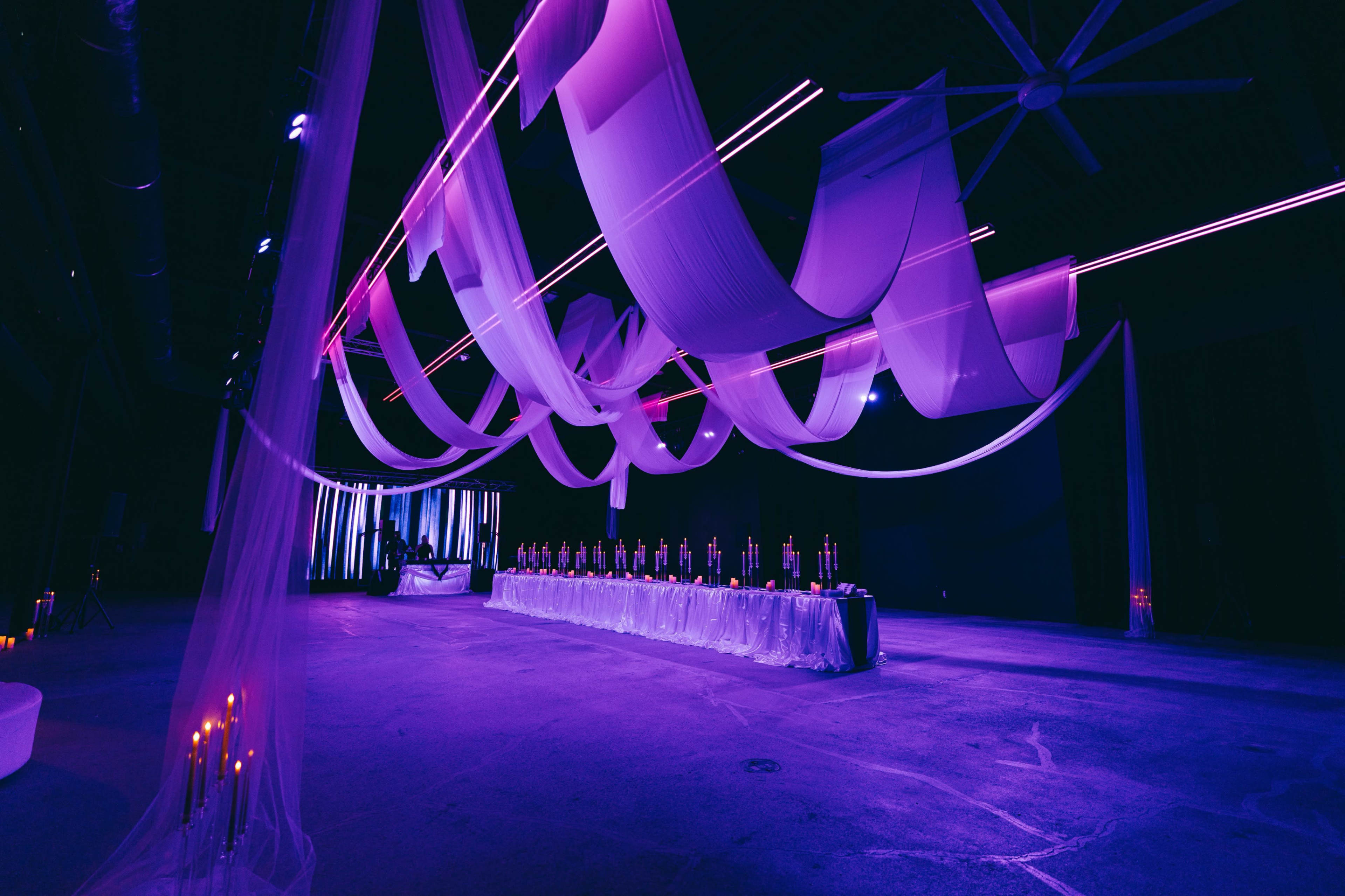 The image shows a large event space decorated with flowing fabric drapes and ambient purple lighting, featuring a long table set with candles at one end.