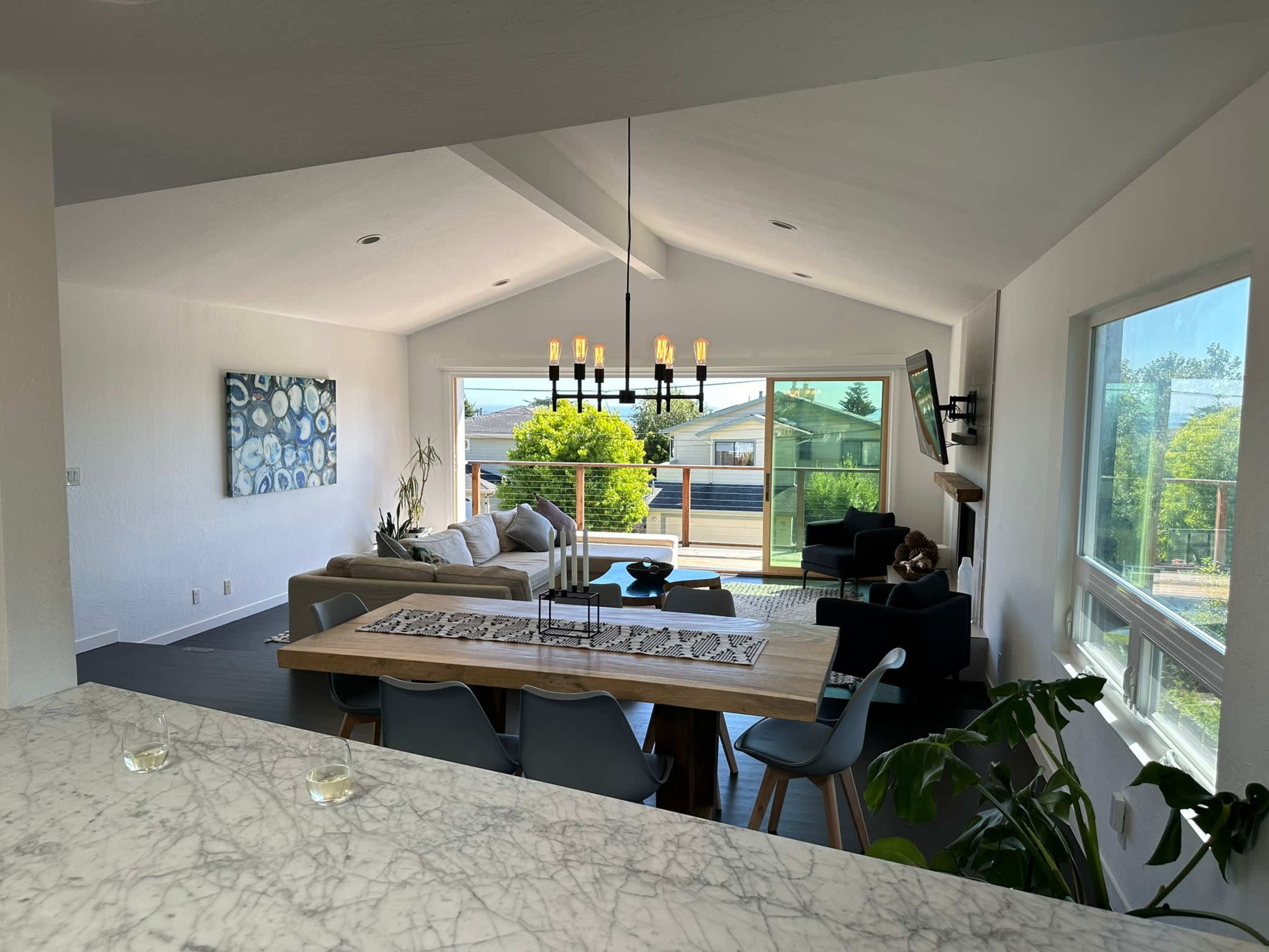 The image shows a modern living room with a dining area, featuring large windows that provide a view of the outdoors, a sectional sofa, and a dining table surrounded by chairs.