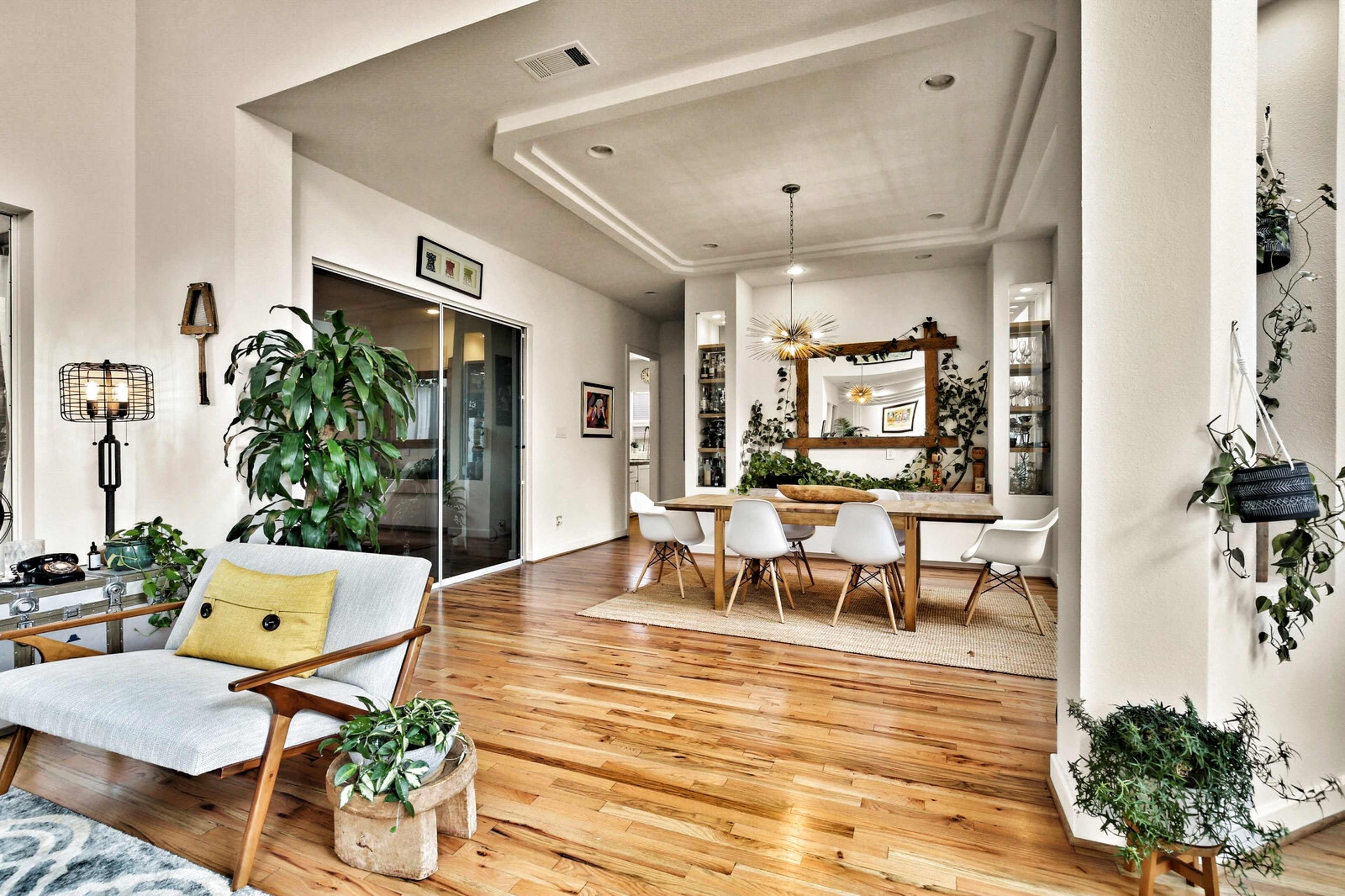A well-lit open-concept living and dining area features wooden floors, a long dining table surrounded by white chairs, and plants decorating the space.