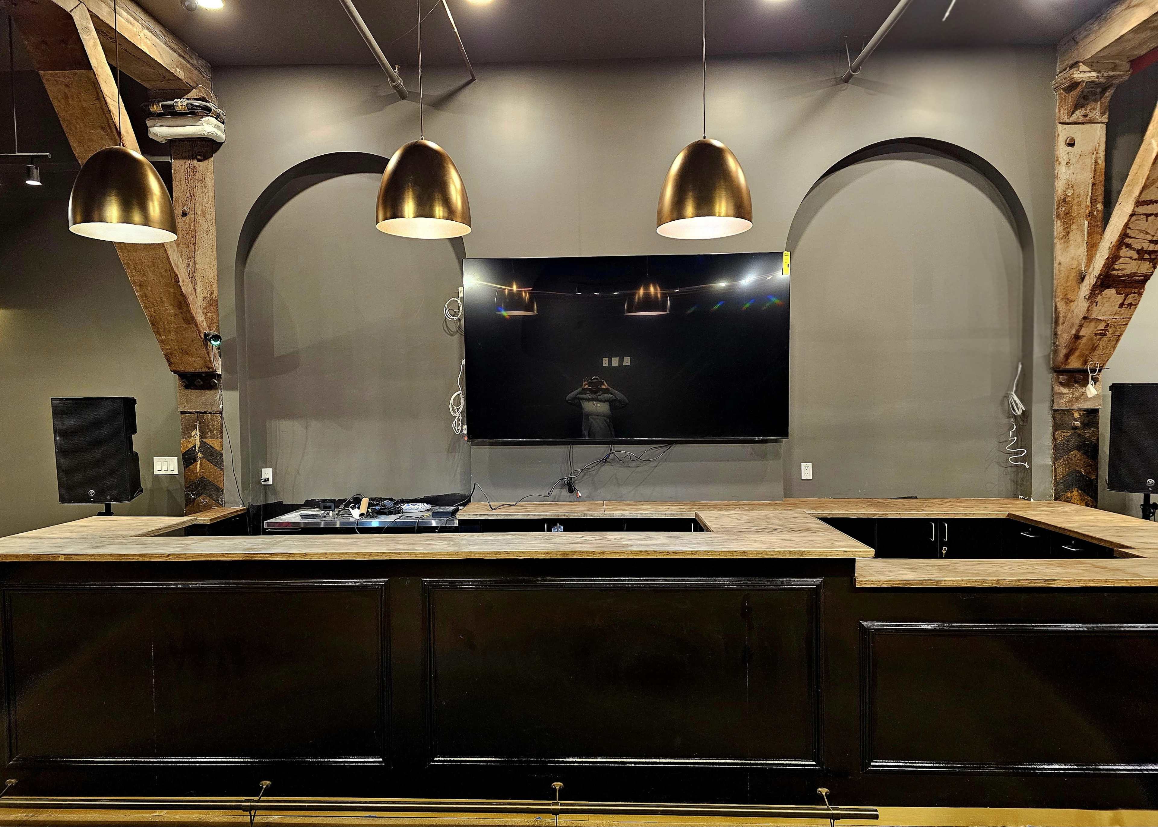 The image shows a bar area featuring a large countertop, two gold pendant lights, and a wall-mounted television, all set against a backdrop of dark walls and exposed wooden beams.