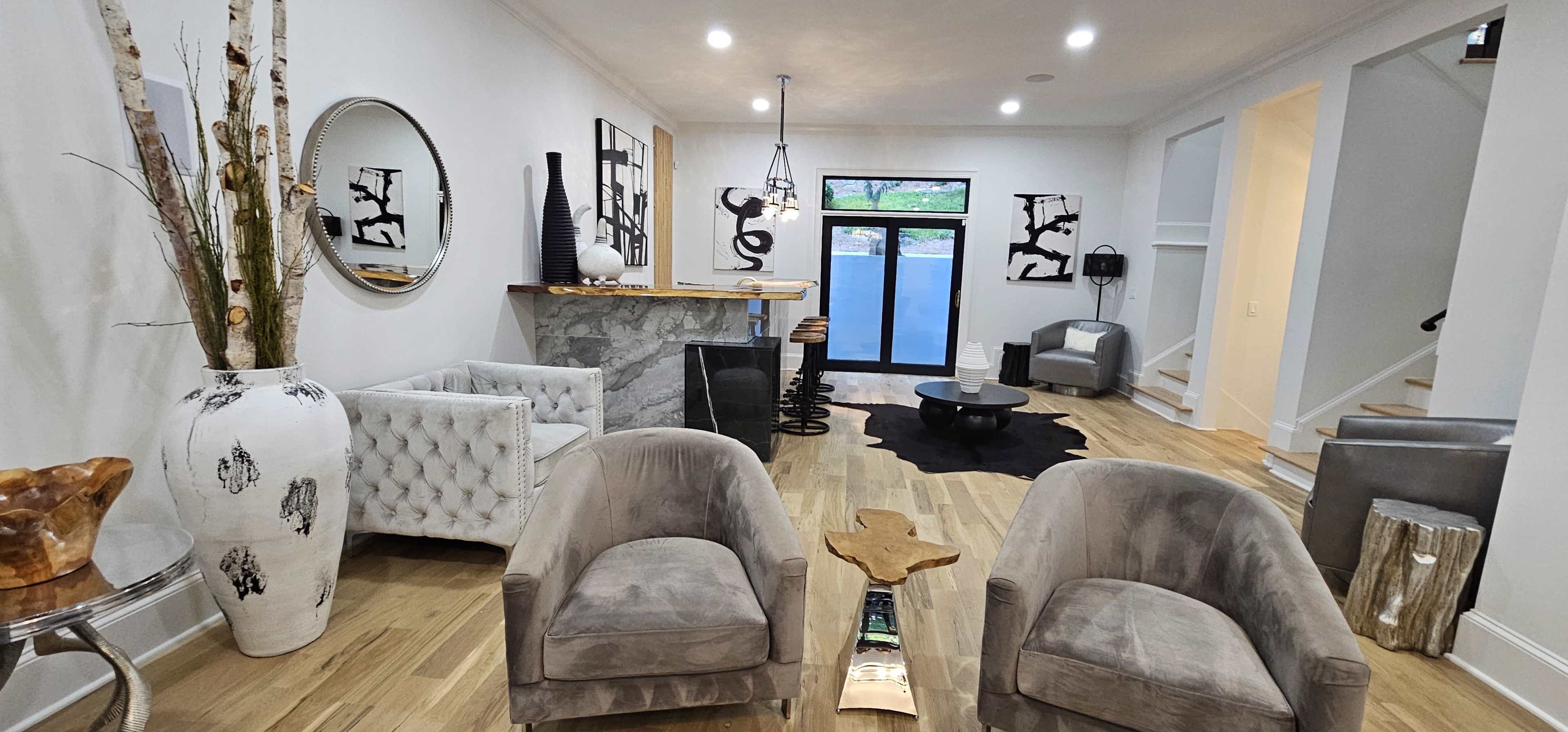 A modern living room with a marble bar, two gray armchairs, a circular mirror, and black and white artwork on the walls.