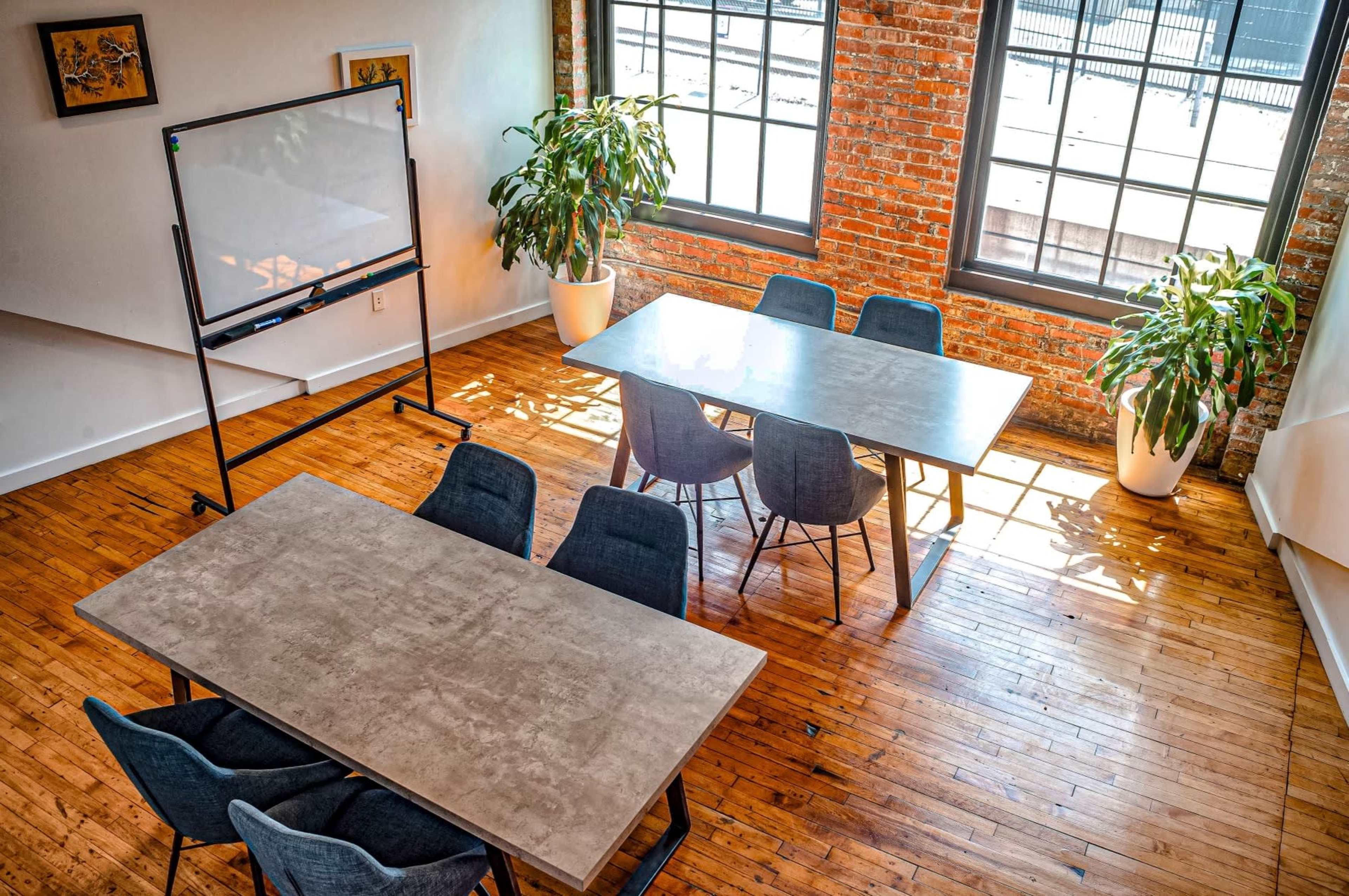 A well-lit conference room featuring two large tables surrounded by gray chairs, a whiteboard on a stand, and potted plants near the windows with exposed brick walls.