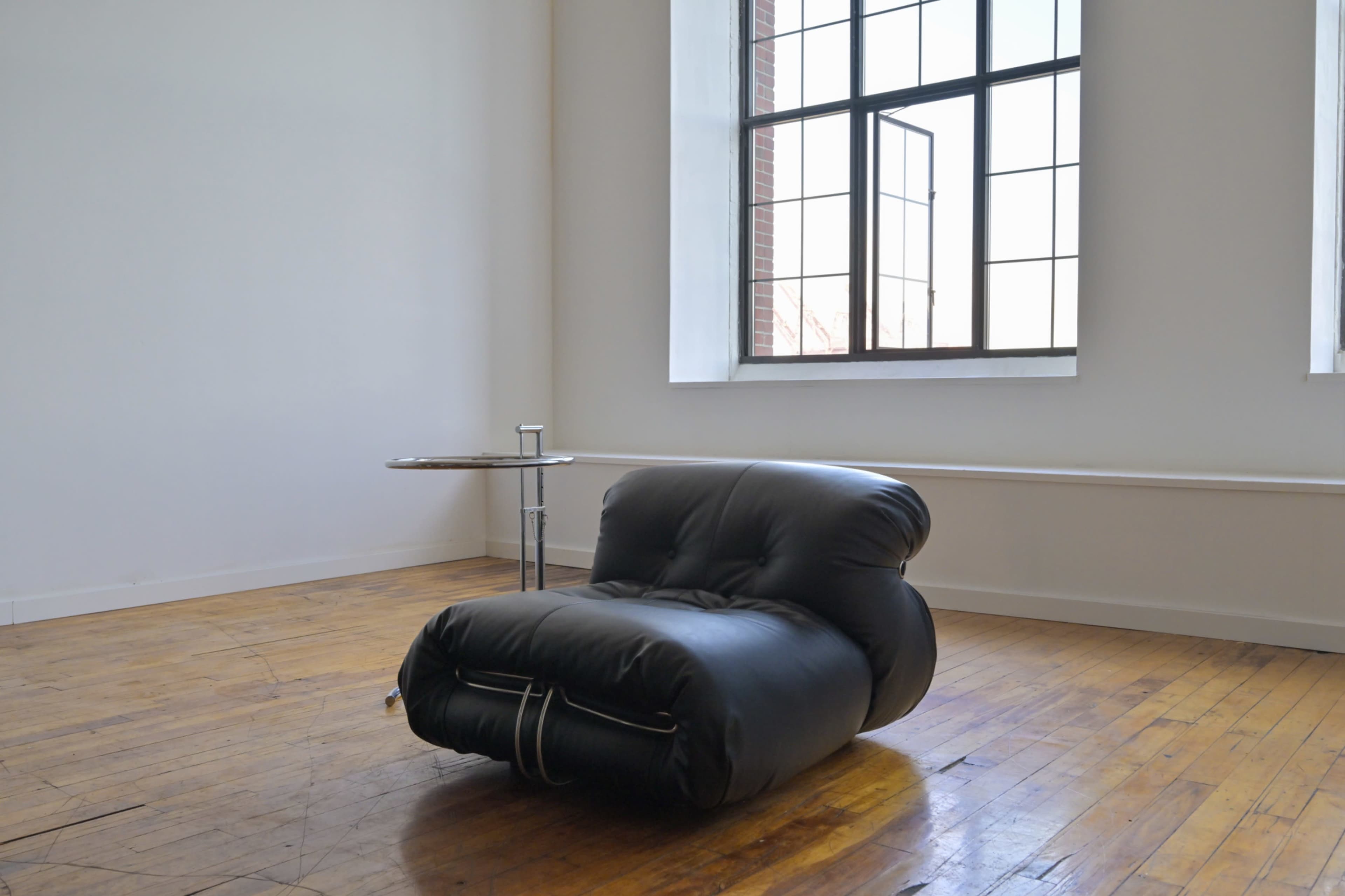 A black, puffy lounge chair sits on a wooden floor near a window in a minimalist room.