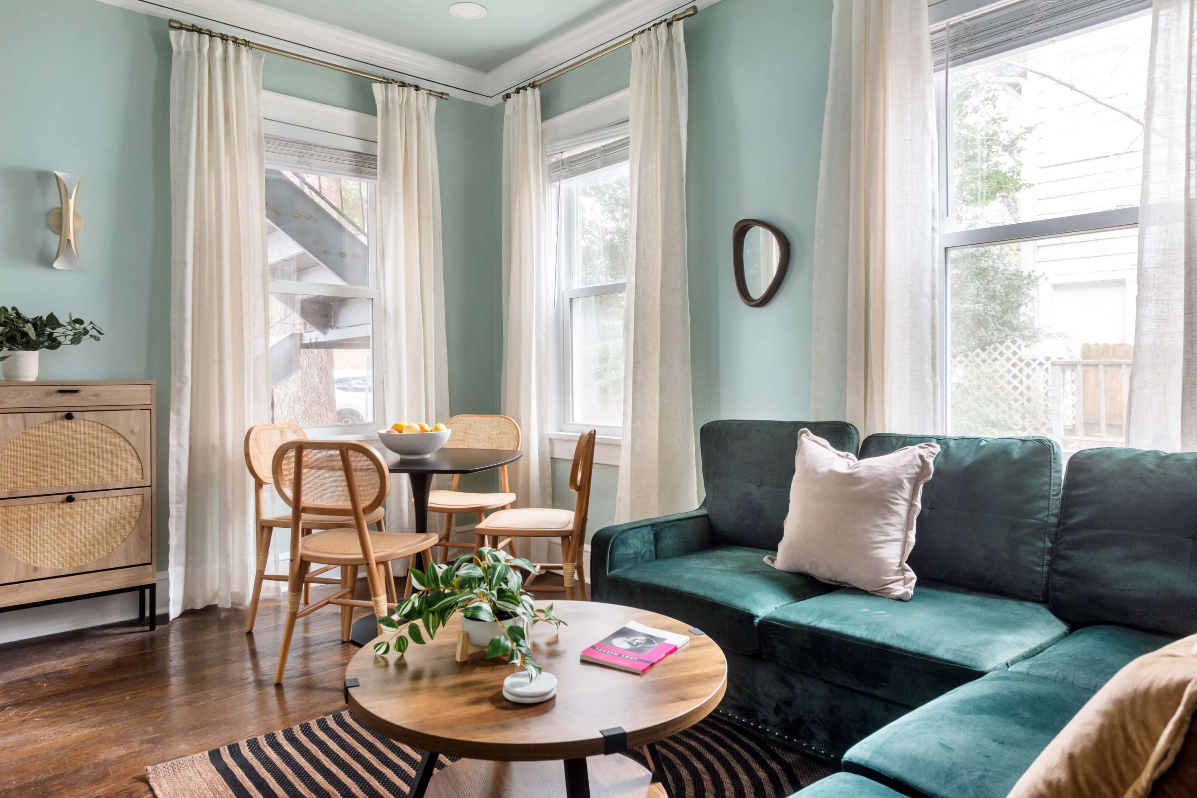 A living room features a green sofa, round wooden coffee table, and a dining area with two chairs and a small table near large windows with light curtains.