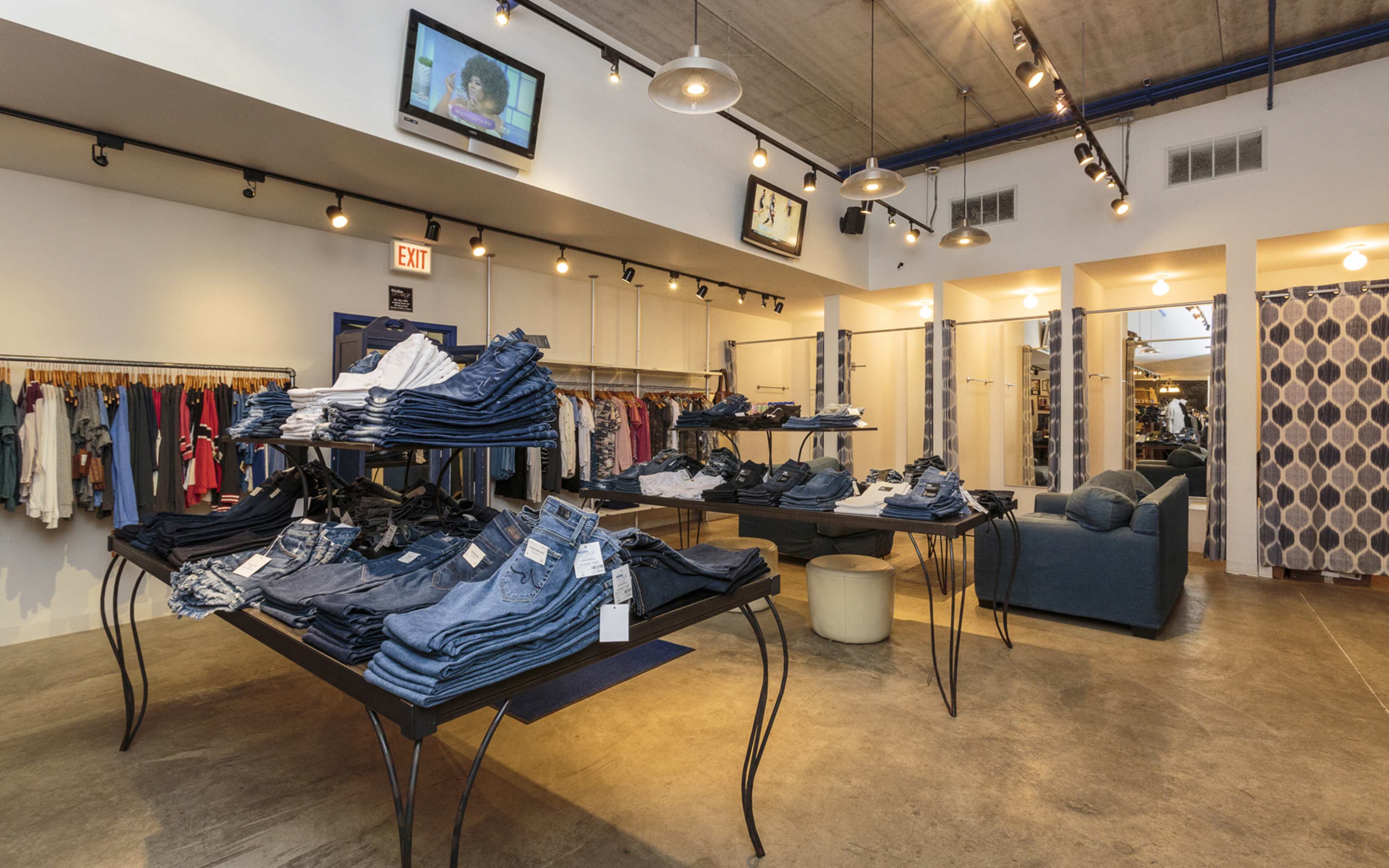 The image shows an interior of a clothing store featuring stacked jeans on tables, clothing racks on the walls, and a television mounted on the ceiling.