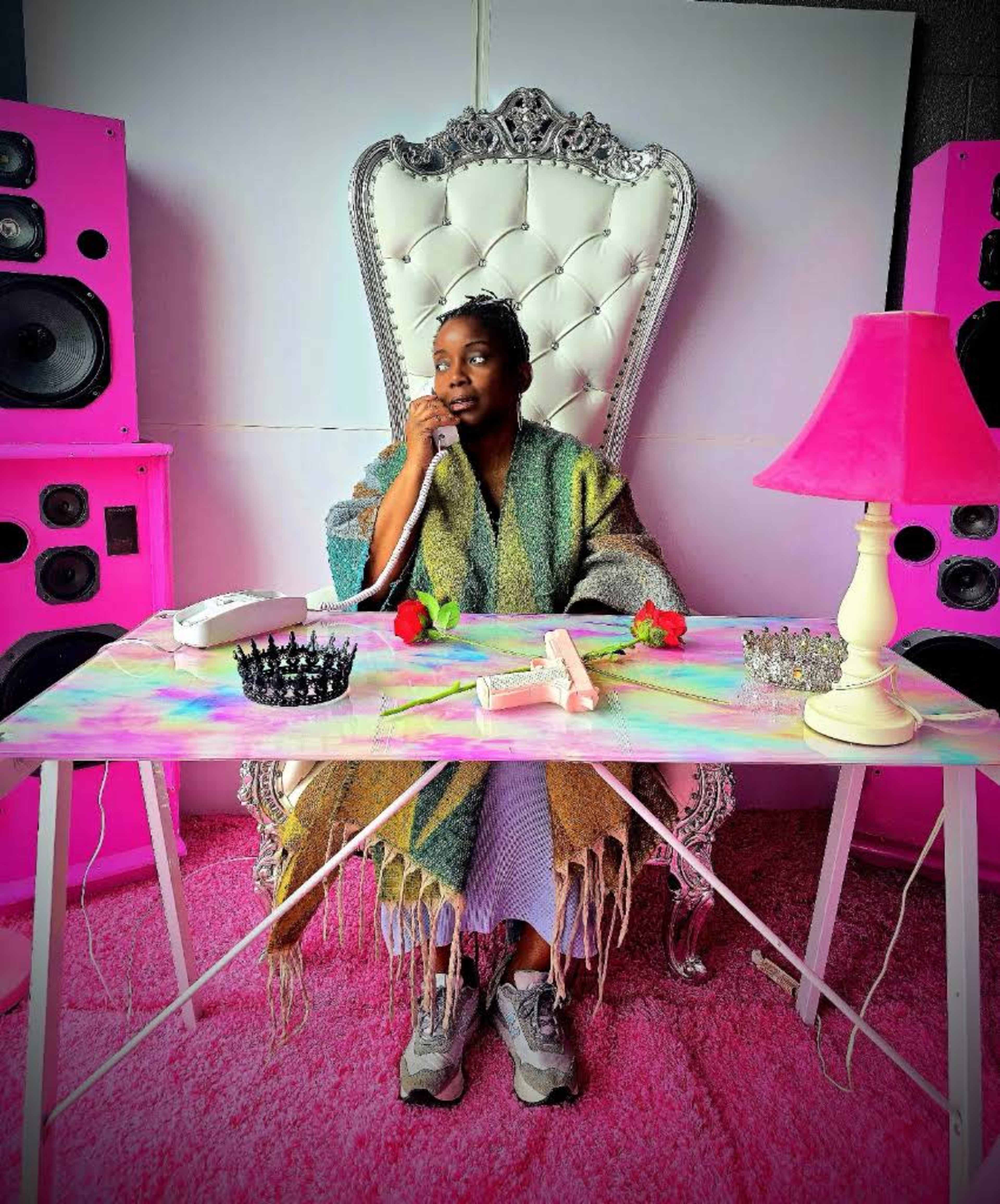 A person sits at a colorful table adorned with flowers and a lamp, speaking on a vintage telephone while flanked by large pink speakers.