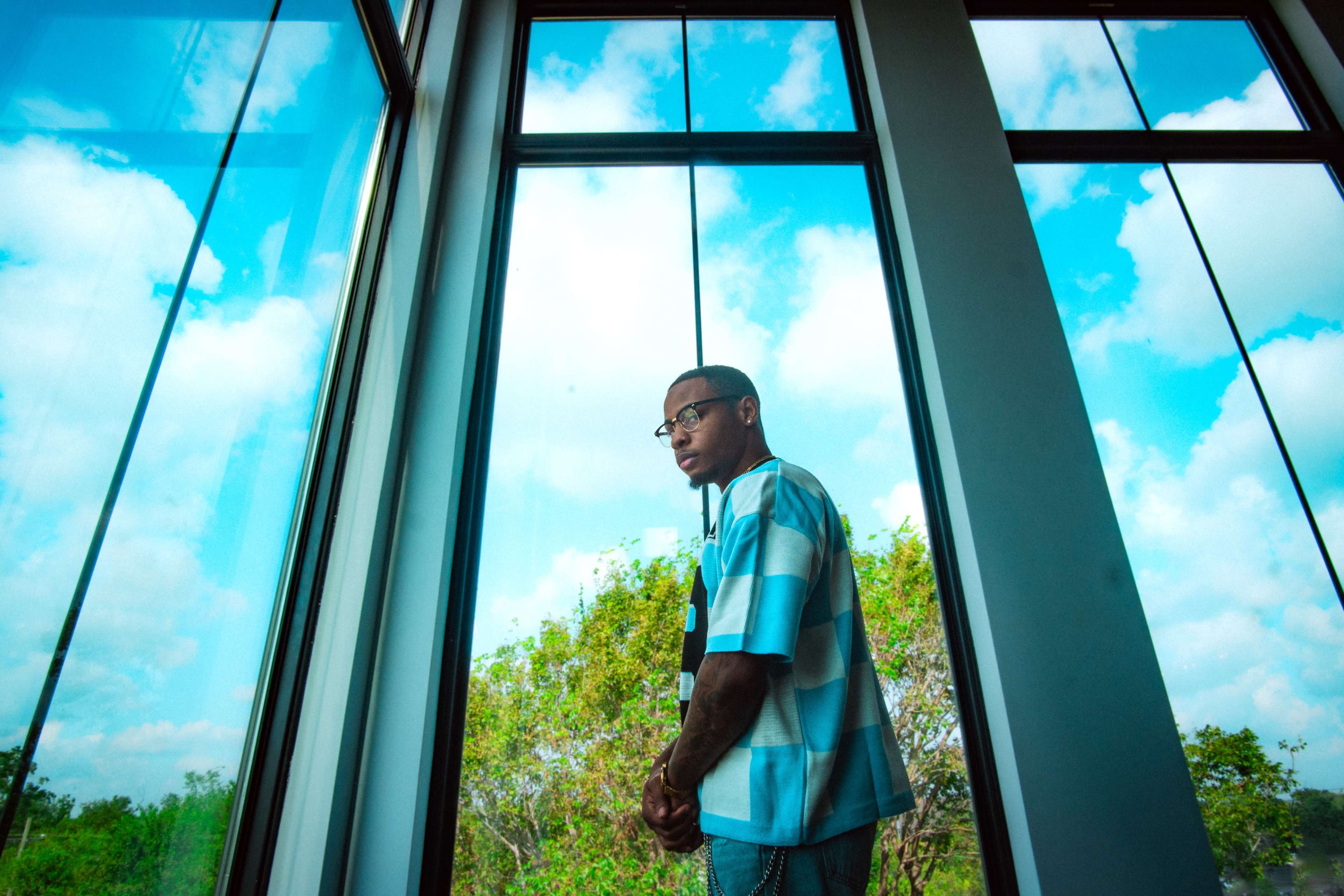 A person stands beside large windows, framed by greenery and a blue sky.