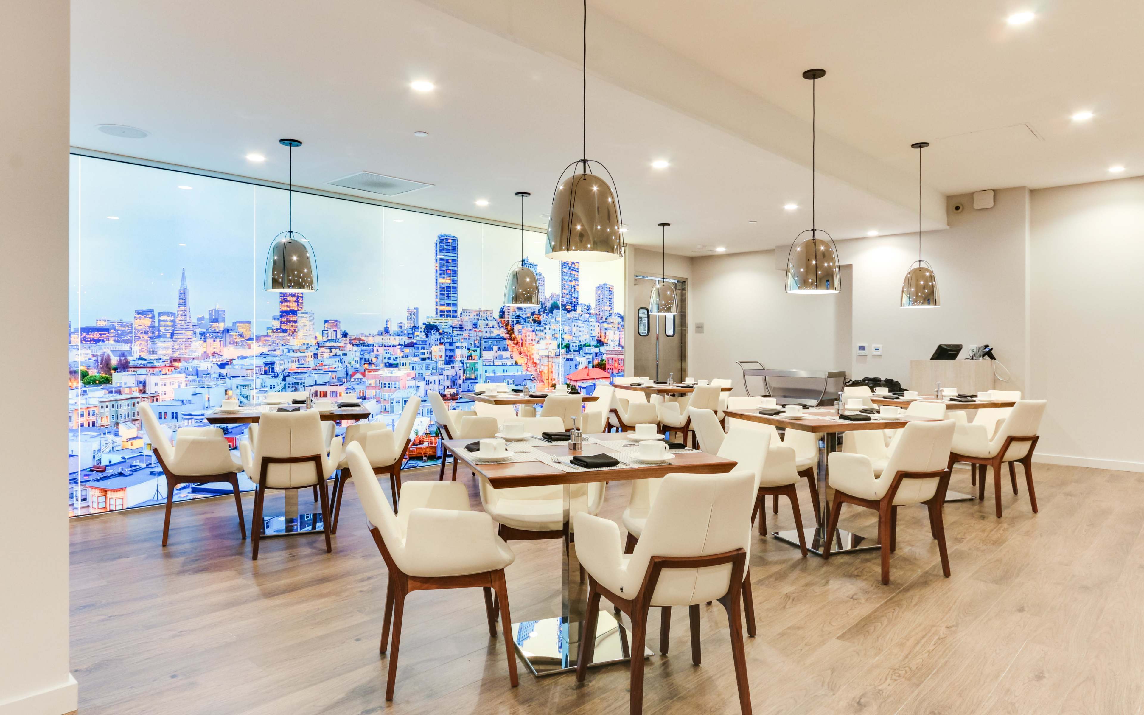 A modern restaurant features white tables and brown chairs, with a large window showcasing a city skyline at dusk.