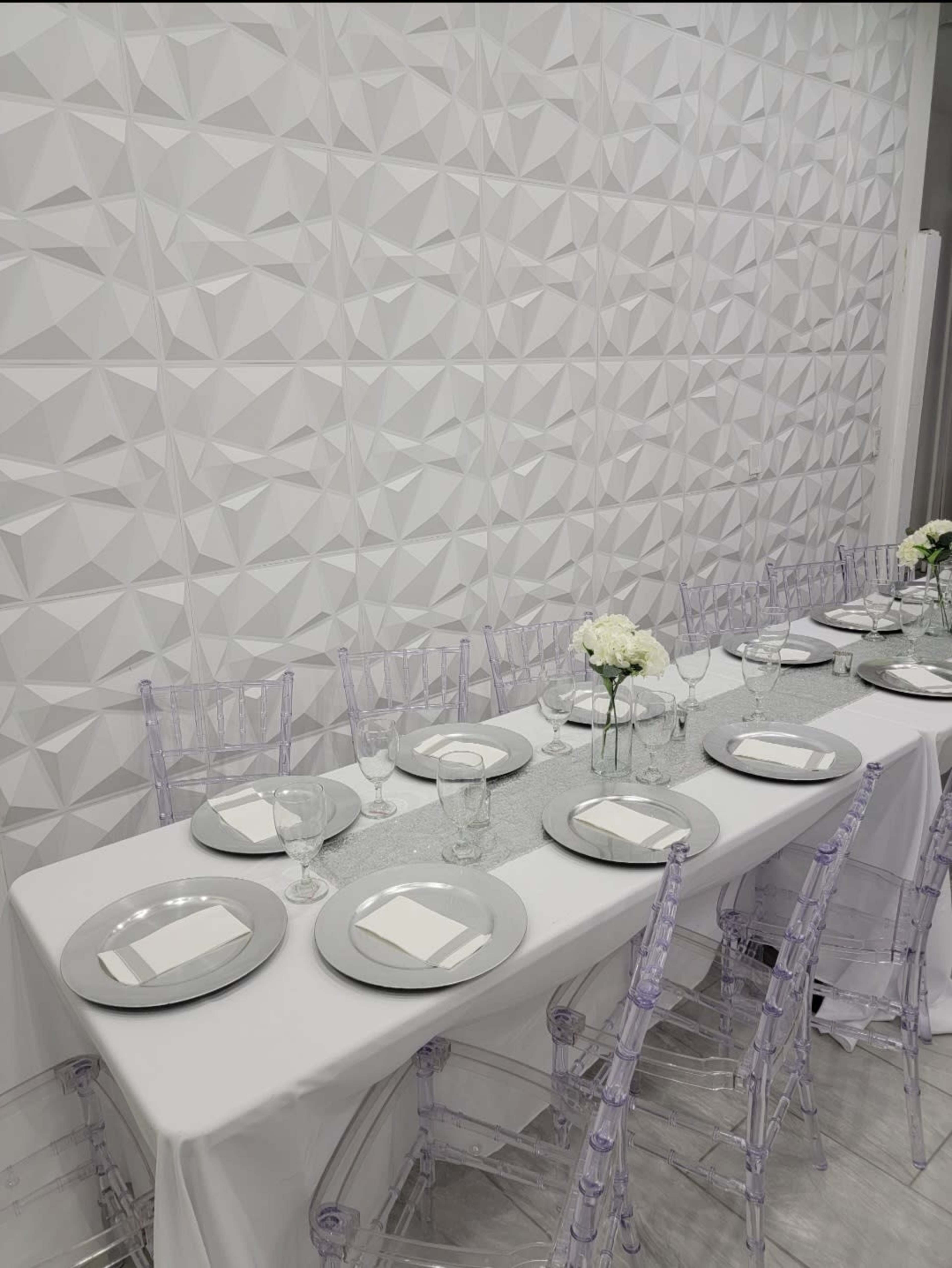 A long table set with silver plates, glassware, and white flowers against a textured white wall.