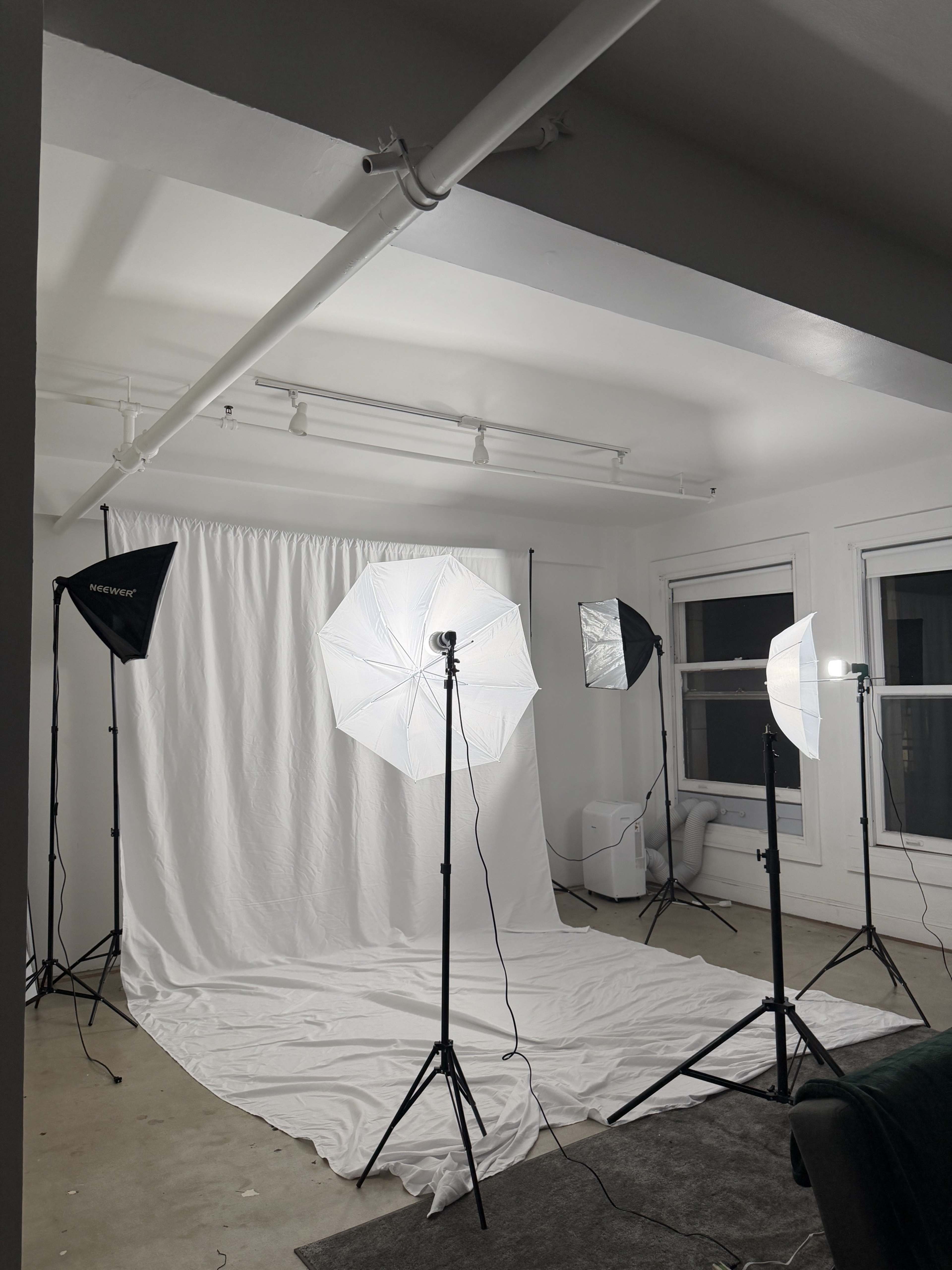 A photography studio setup features multiple softbox lights arranged around a white backdrop and a draped surface on the floor.