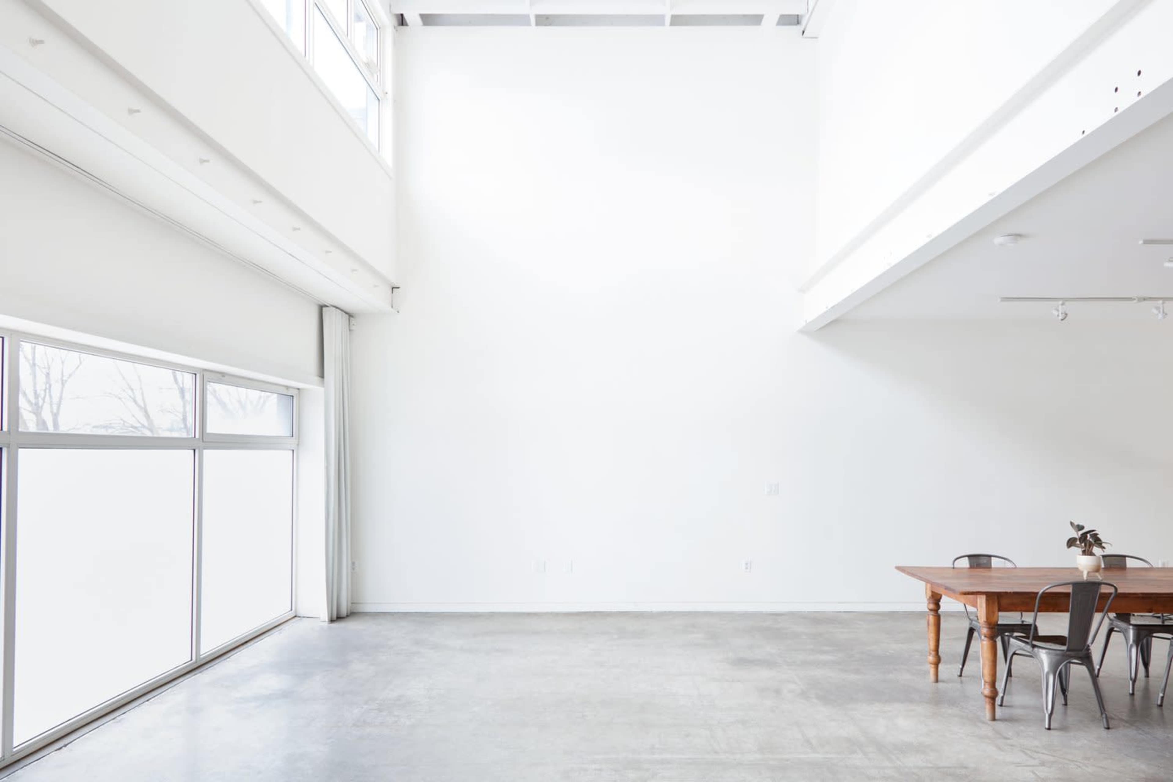 A spacious interior with high white walls, a large window, and a wooden dining table surrounded by chairs.