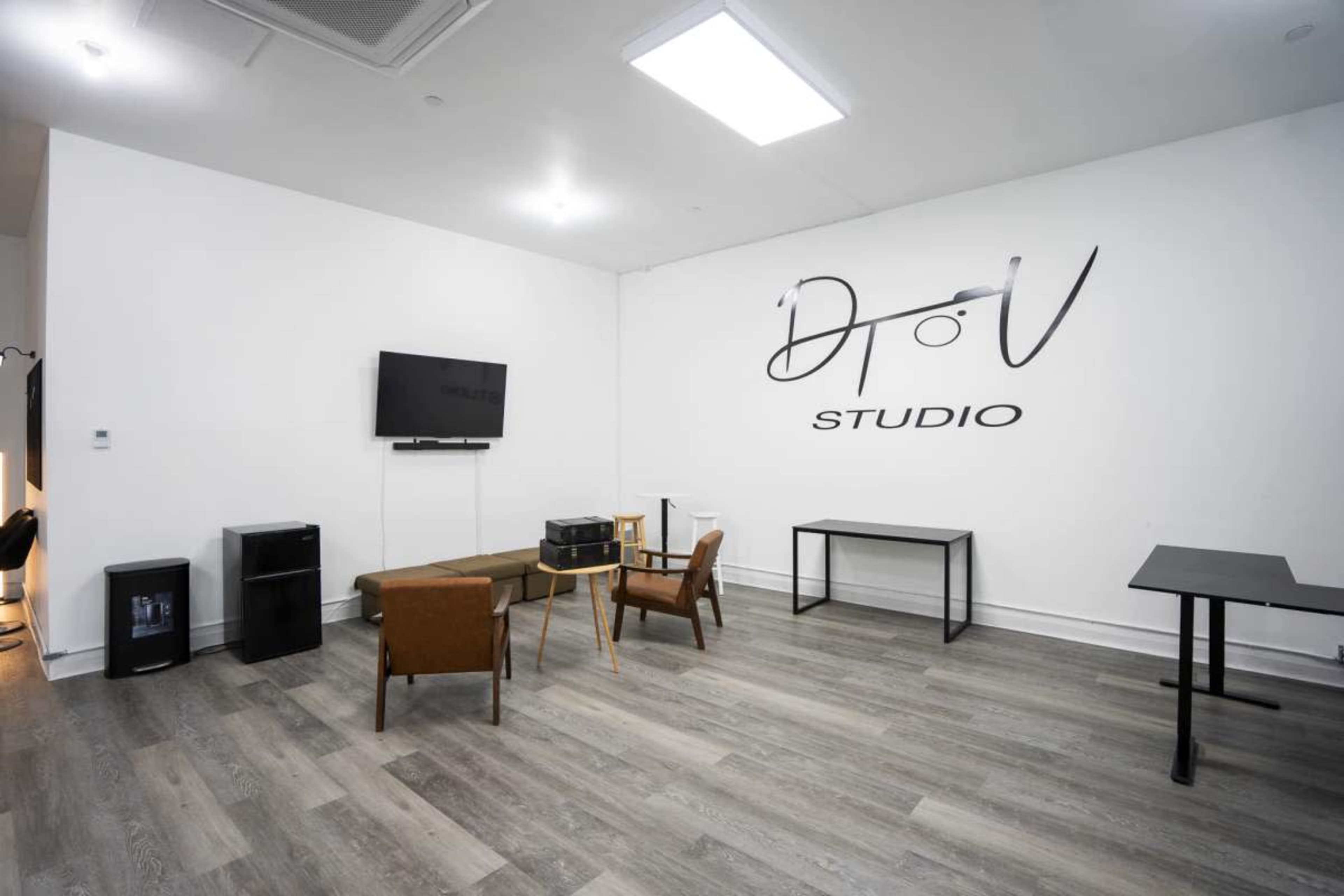 The image shows a modern studio space with a television mounted on the wall, a wooden sofa, two chairs, and a table, all set against a white backdrop featuring the text "DToV STUDIO."