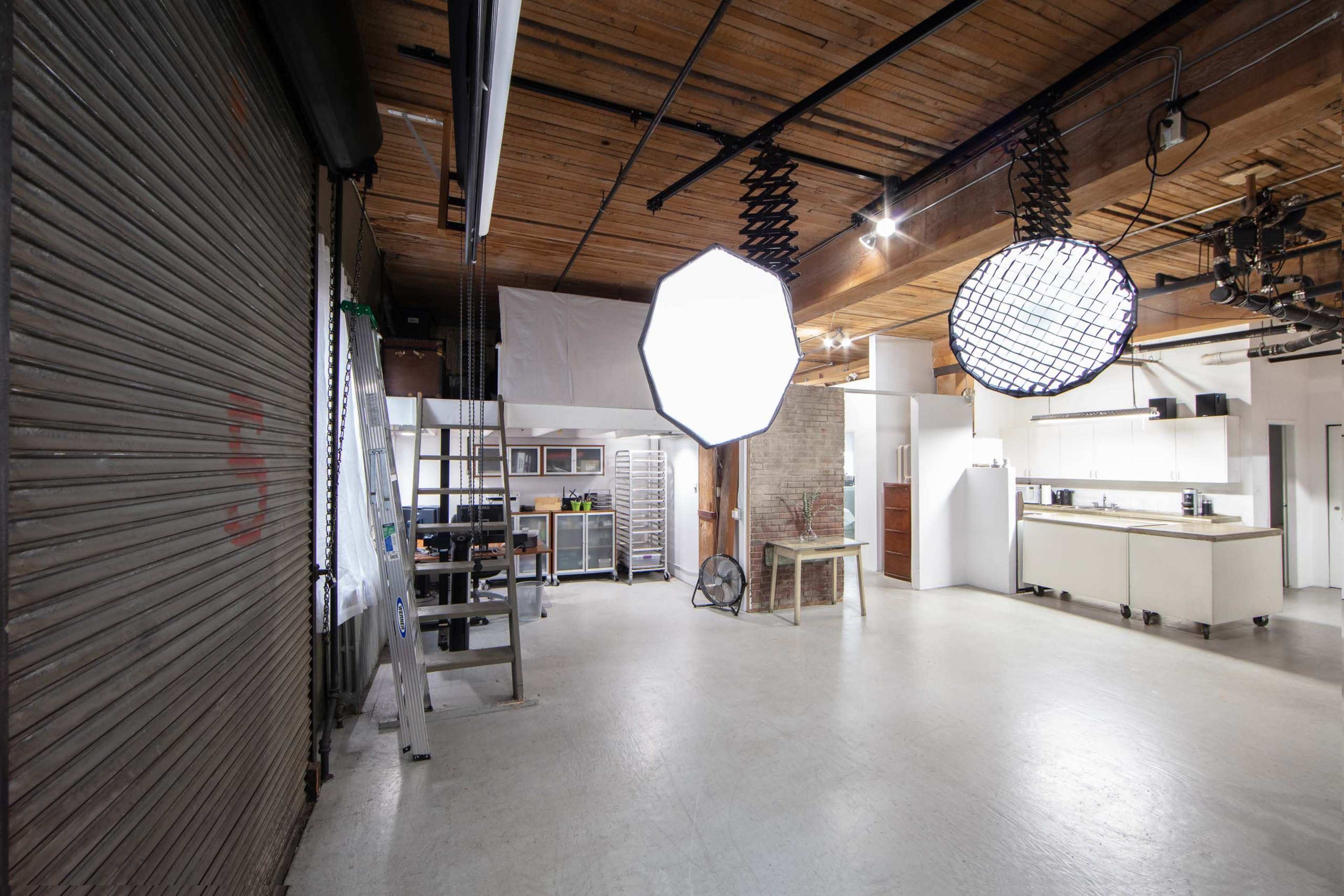 An industrial-style studio space with wooden beams, a large overhead light, and a kitchen area in the background.