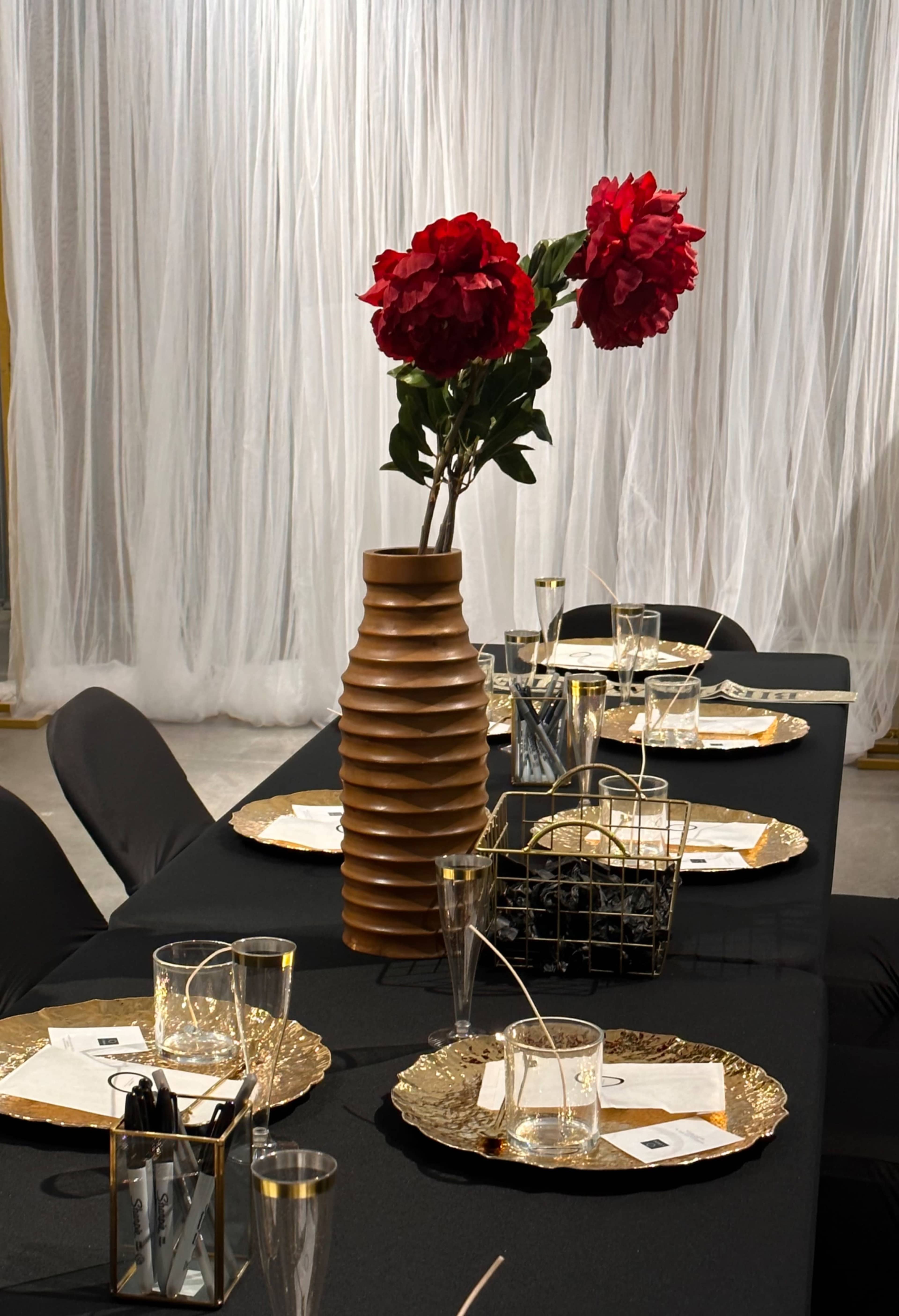 A long dining table is set with gold-rimmed plates and clear glasses, featuring a tall vase with red flowers as the centerpiece.