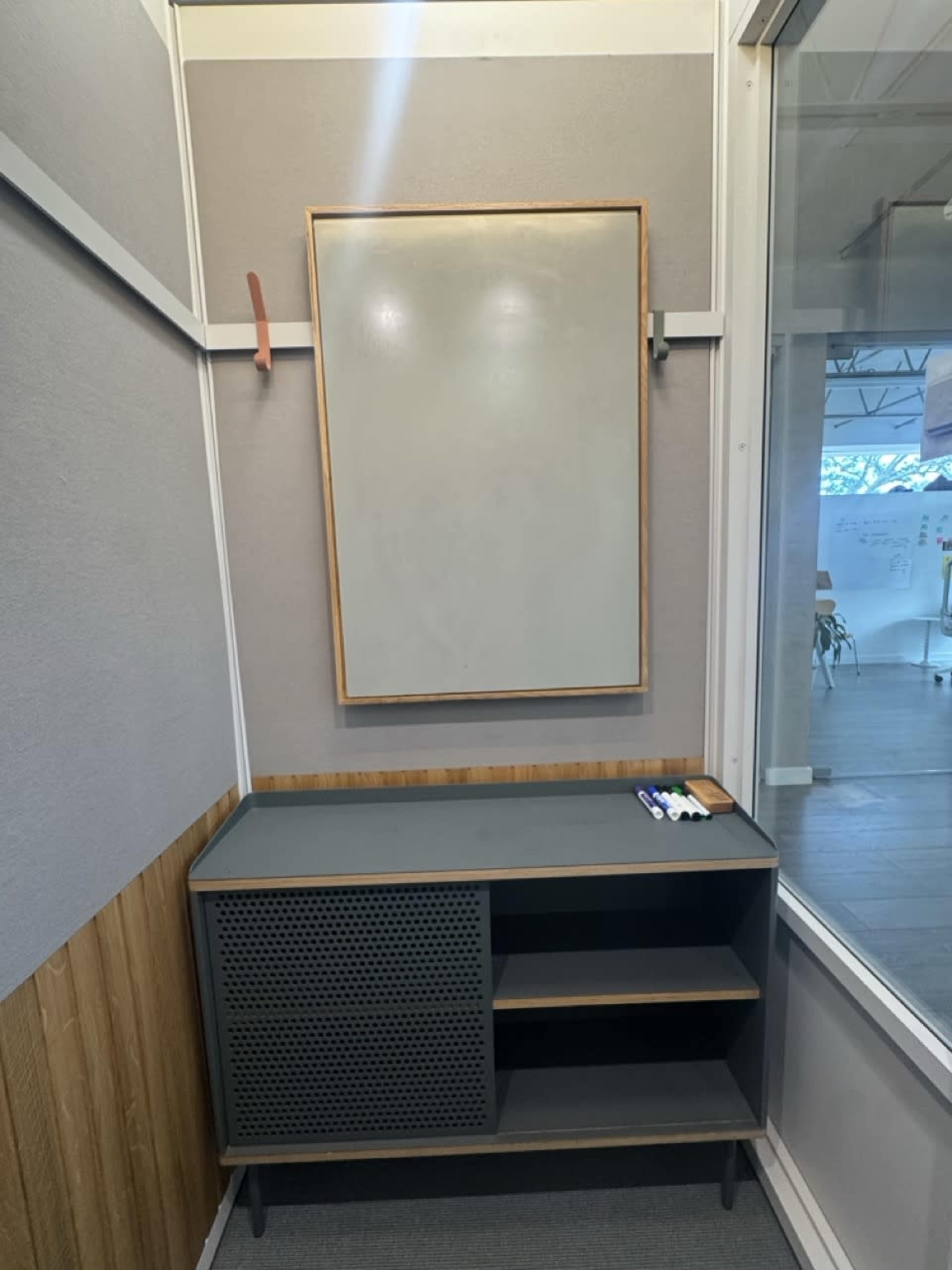 The image shows a minimalist corner of a room featuring a wall-mounted whiteboard, a framed mirror, and a modern gray cabinet with open shelves and a perforated front.
