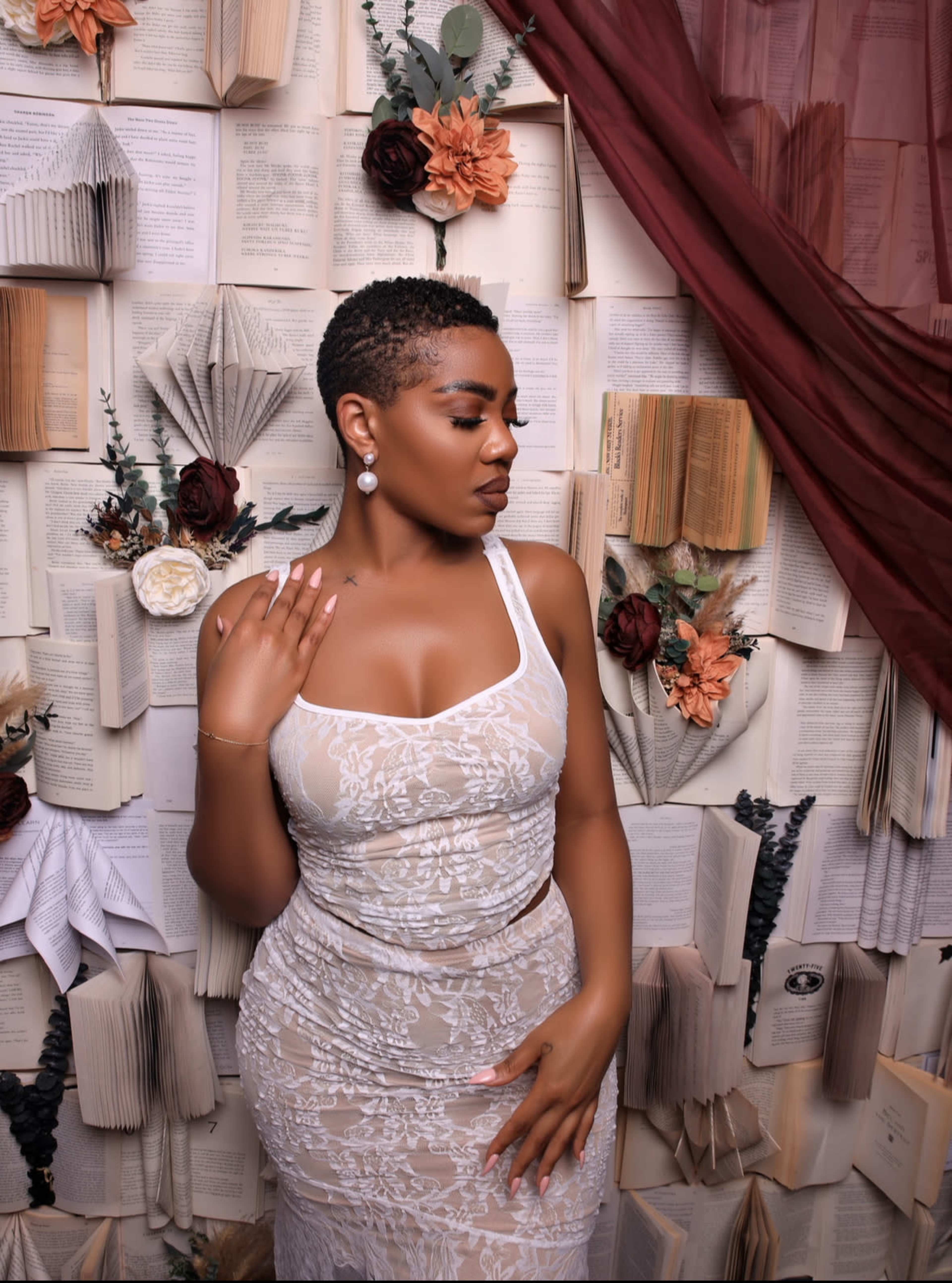 A woman in a fitted, sheer dress poses in front of a backdrop made of open books and floral elements.