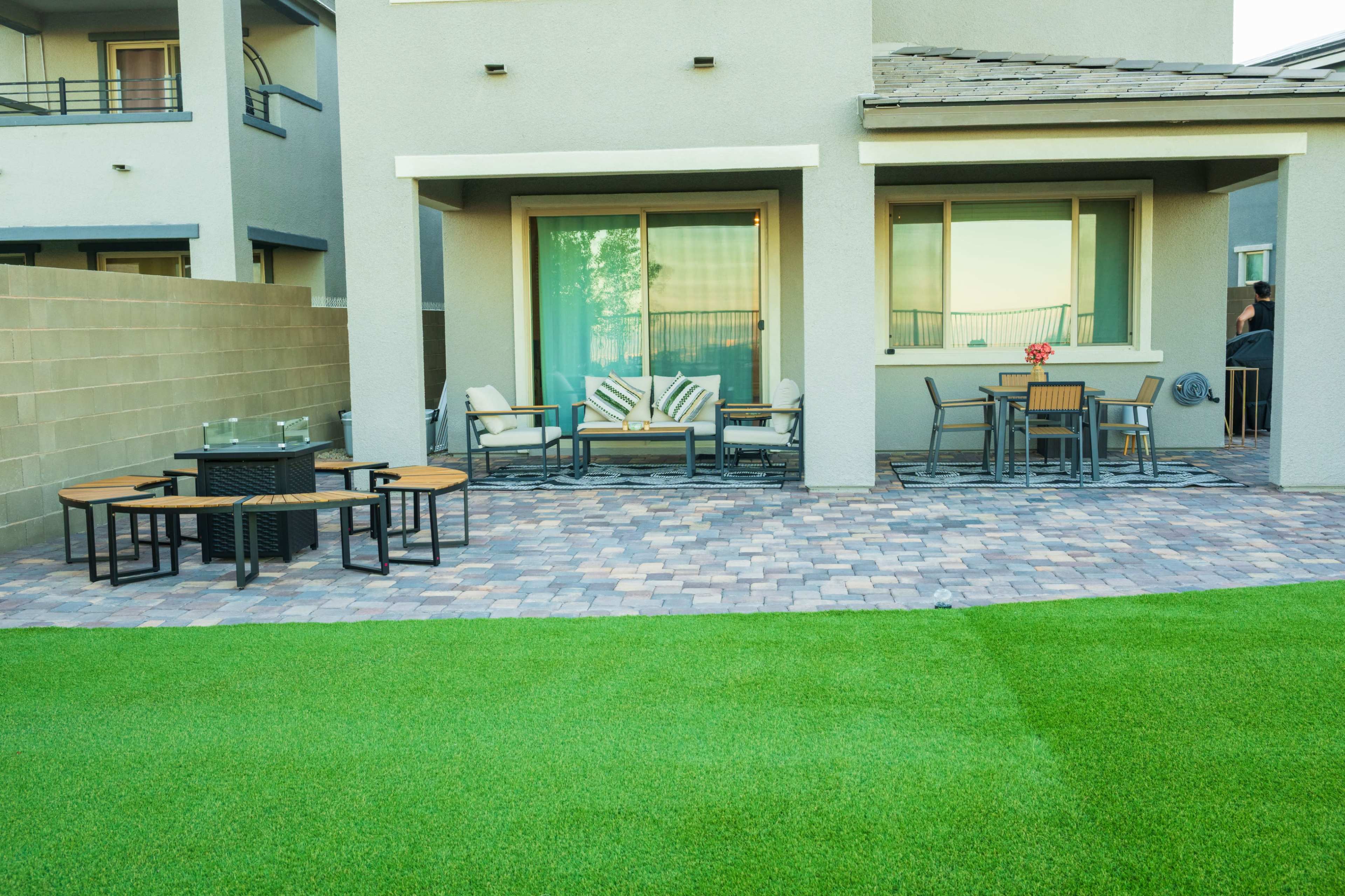 A patio area features a seating arrangement with a table and chairs, surrounded by a manicured lawn and a stone walkway.