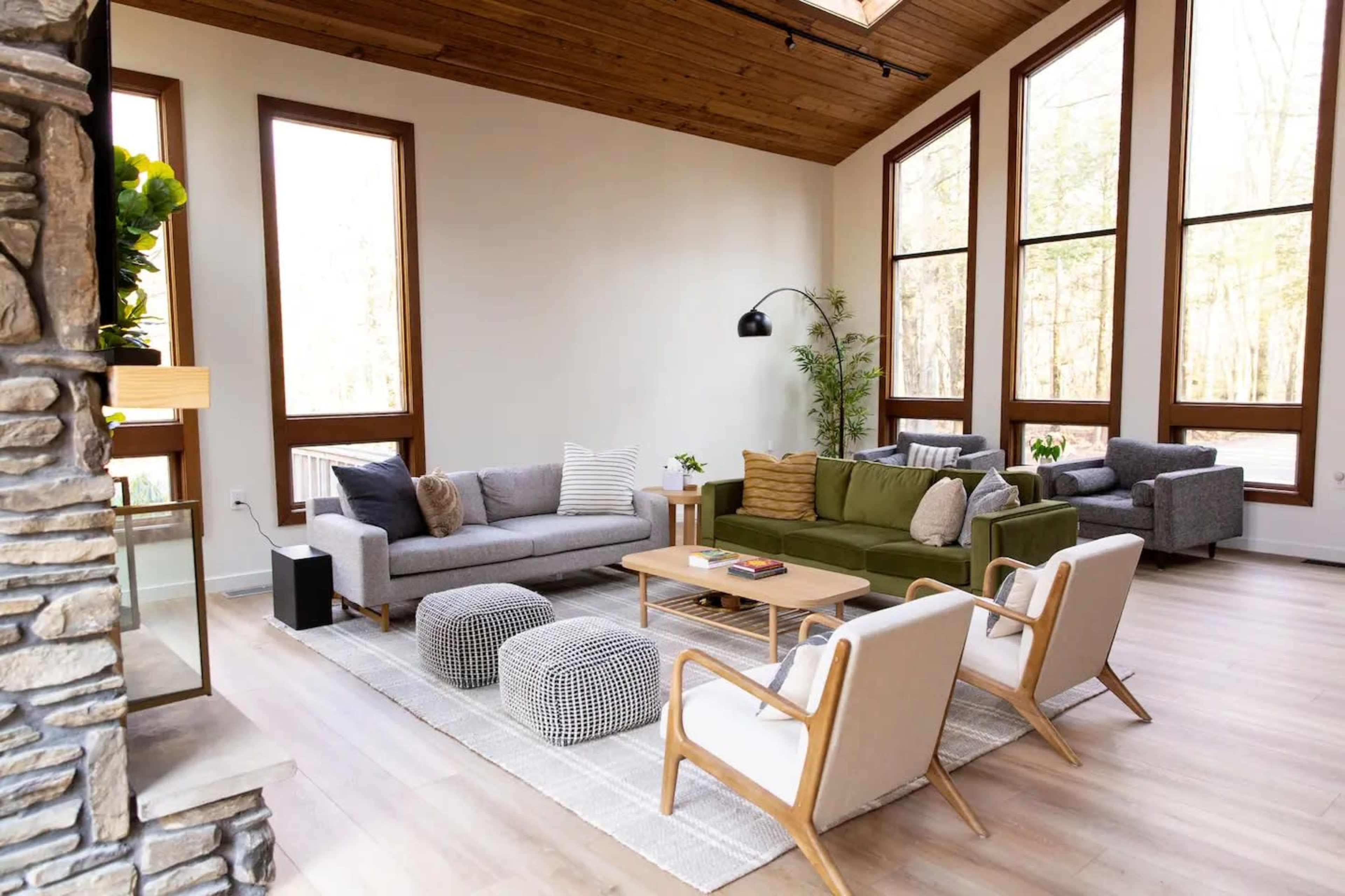 The image shows a spacious, modern living room with large windows, a stone fireplace, and various seating arrangements including sofas and armchairs.