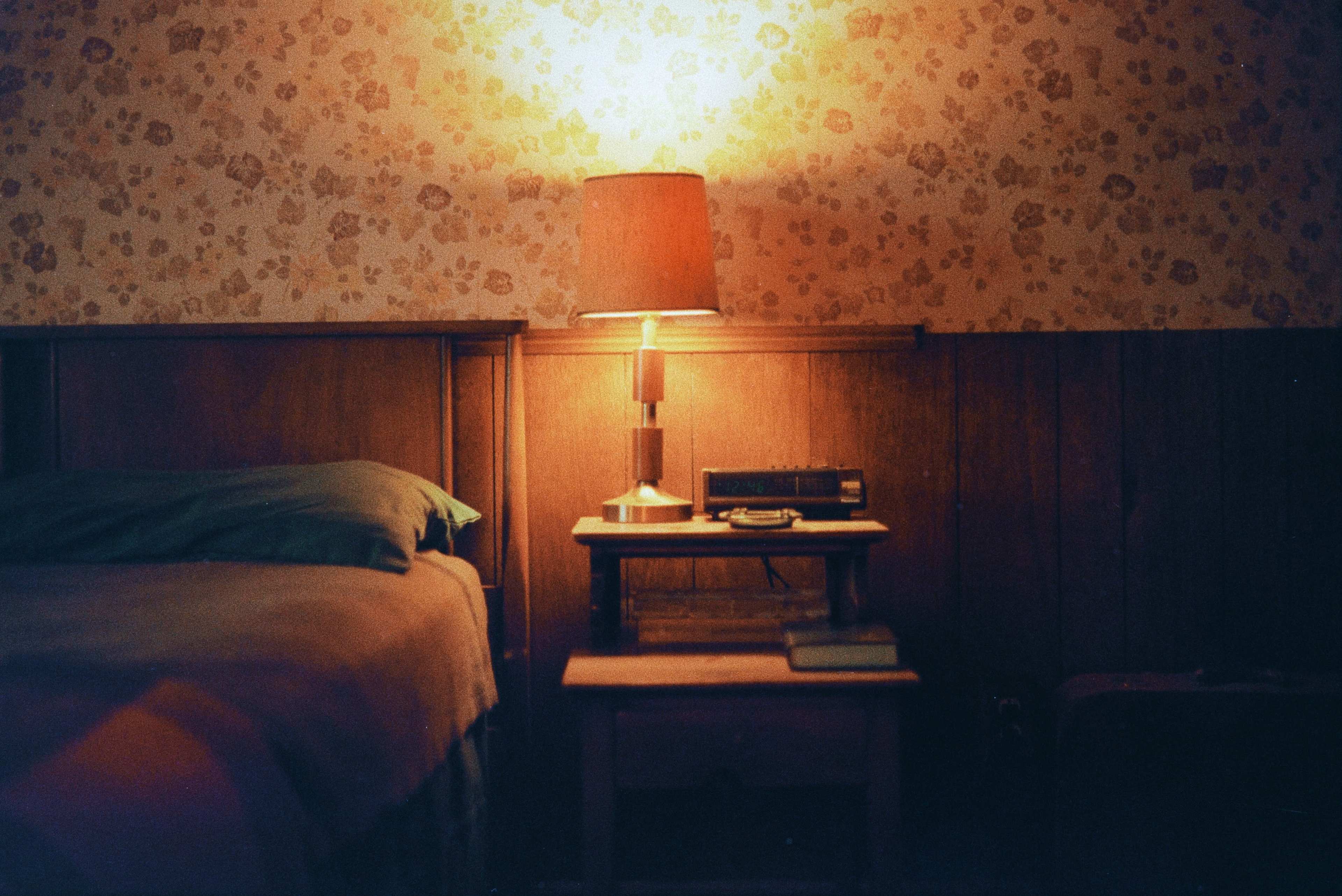 A bedside table with a lamp, clock radio, and a book sits next to a bed in a room with patterned wallpaper.