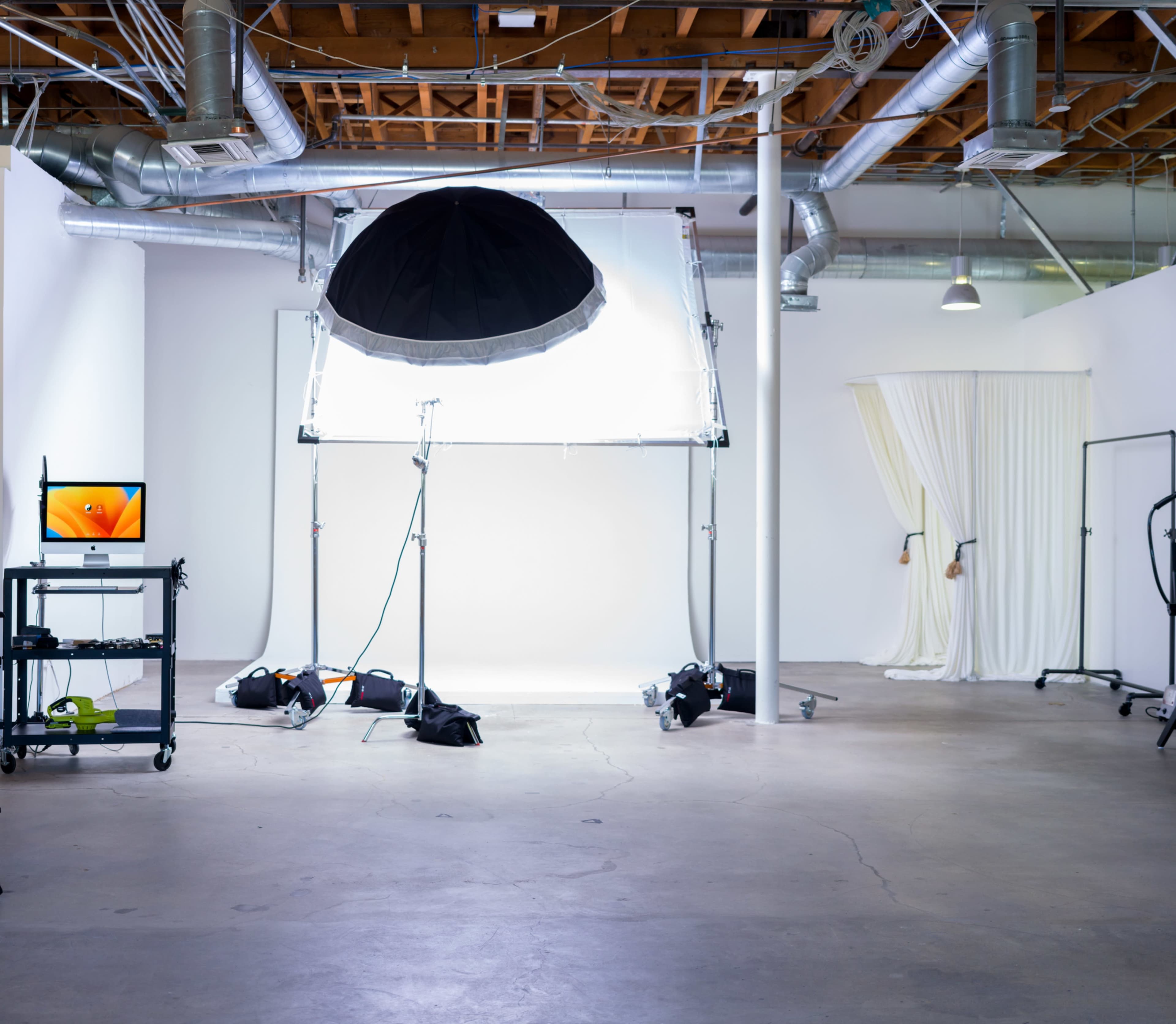 A photography studio with a large softbox light setup, a monitor on a cart, and a backdrop featuring draped fabric.