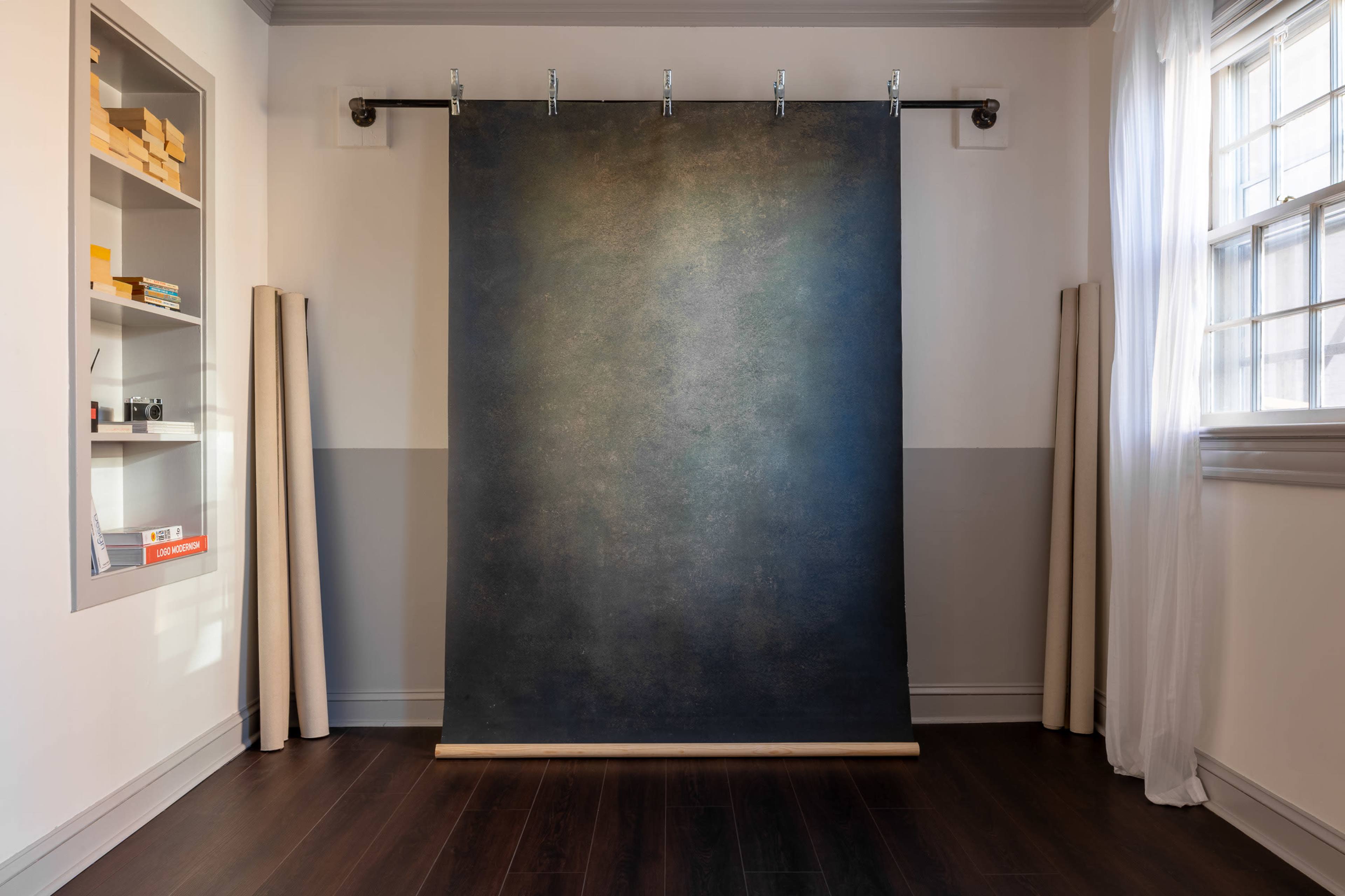 The image shows a photography backdrop hung on a wooden rod in a well-lit room with dark wooden flooring and shelves containing various items.