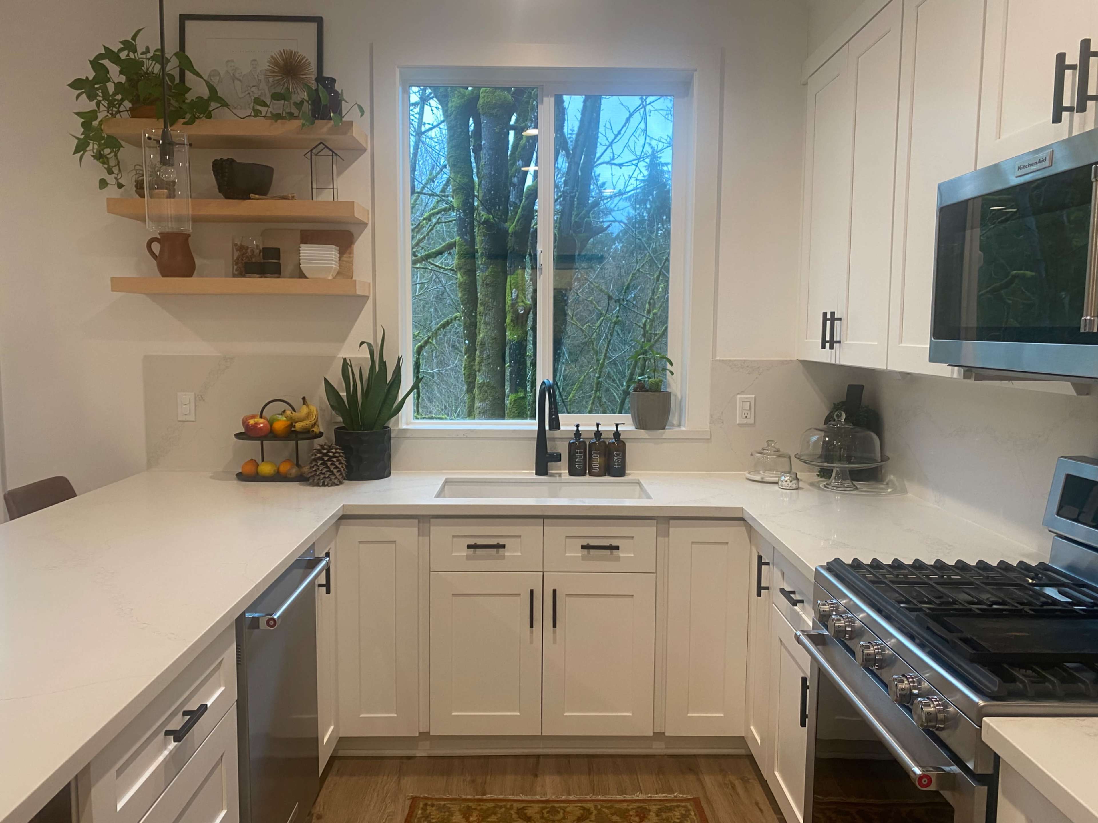 A modern kitchen features white cabinetry, stainless steel appliances, and a window overlooking a wooded area.