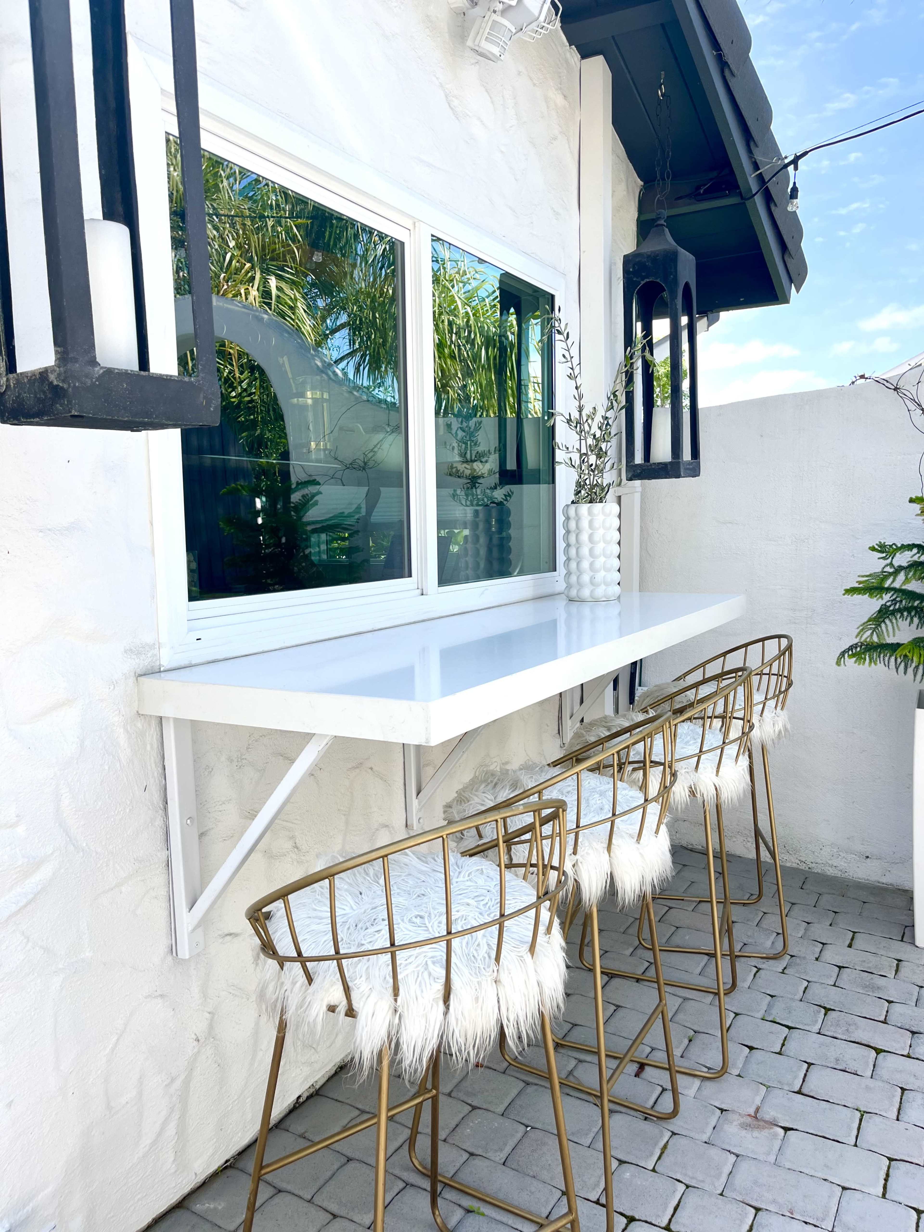 A white outdoor countertop is positioned against a wall, featuring three gold wire stools with furry white cushions.