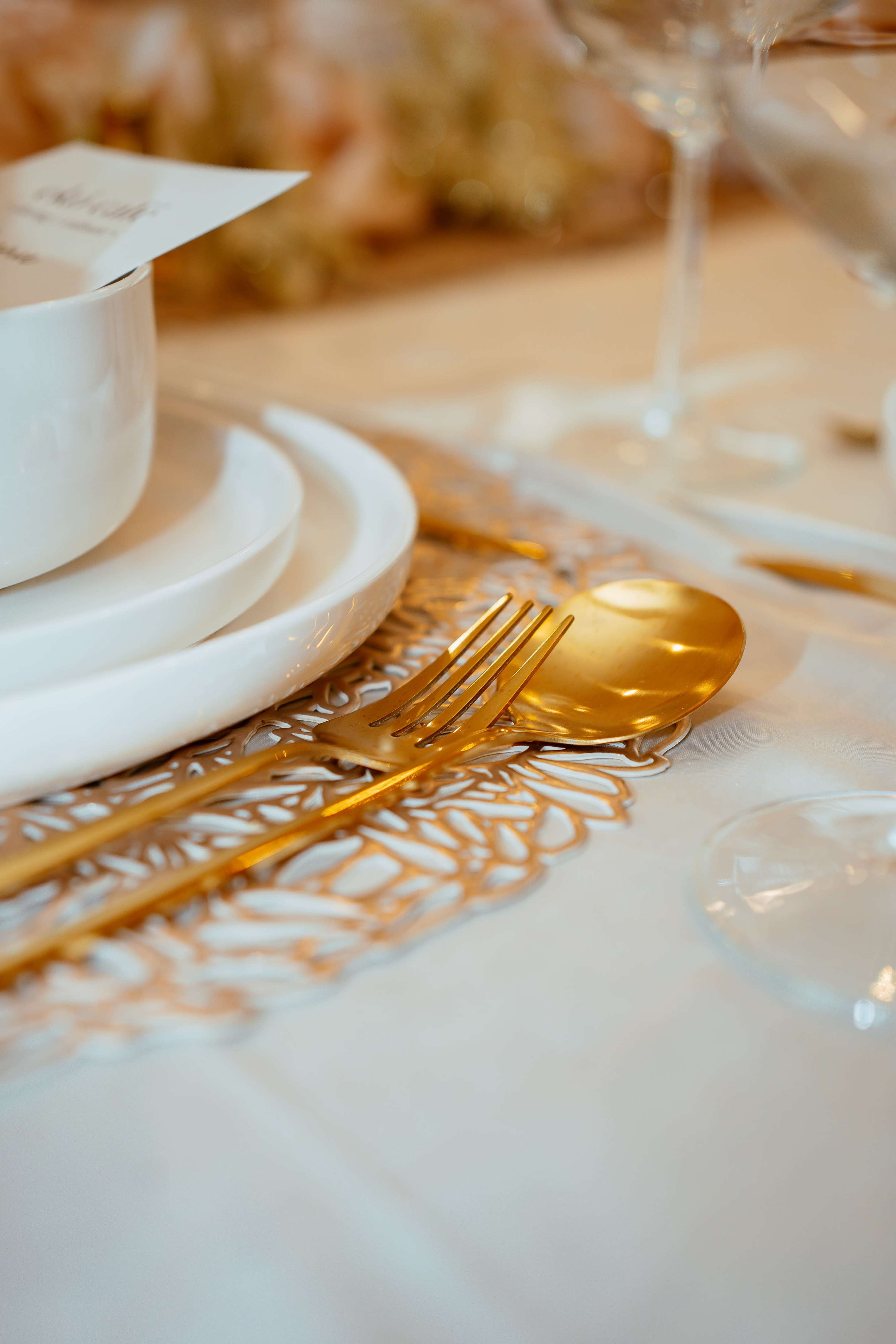 The image features a elegantly set dining table with white dinnerware, a decorative gold-colored placemat, and golden cutlery.