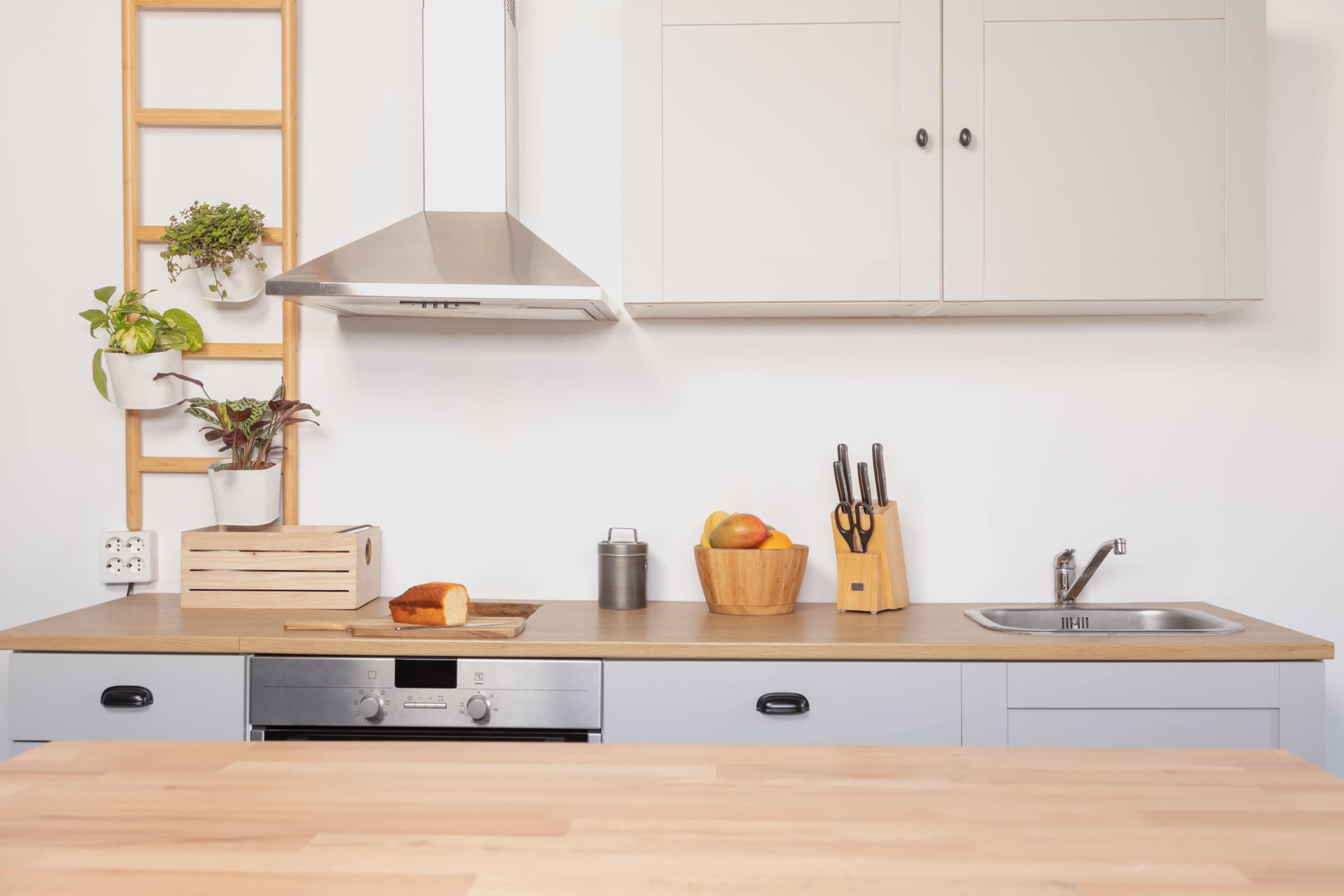 A modern kitchen features a wooden countertop with a loaf of bread, a metal canister, a bowl of fruit, a knife block, and a stainless steel sink against light-colored cabinets and a silver range hood.