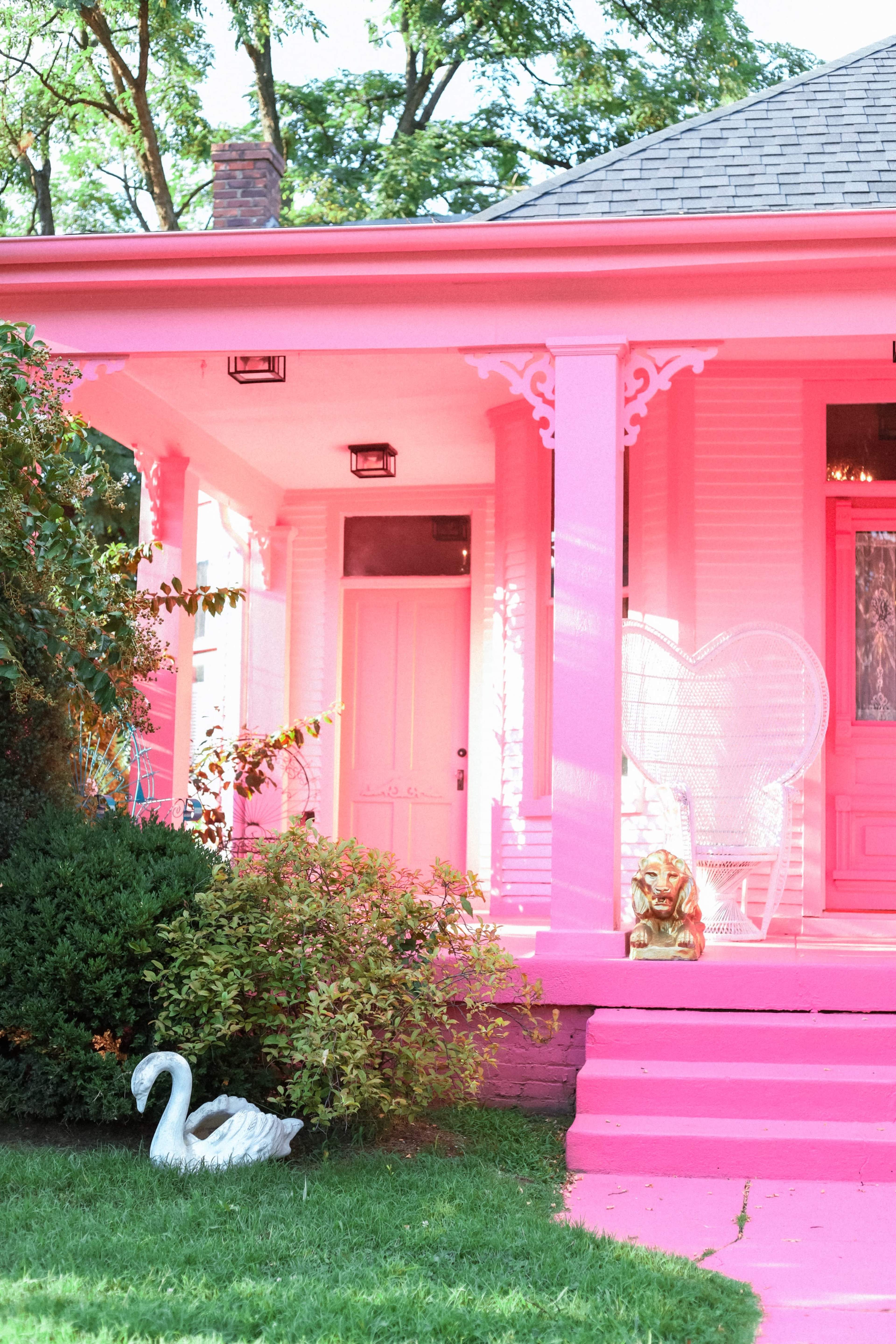 The image shows a vibrant pink house with a matching front porch and a decorative white swan statue in the grassy yard.
