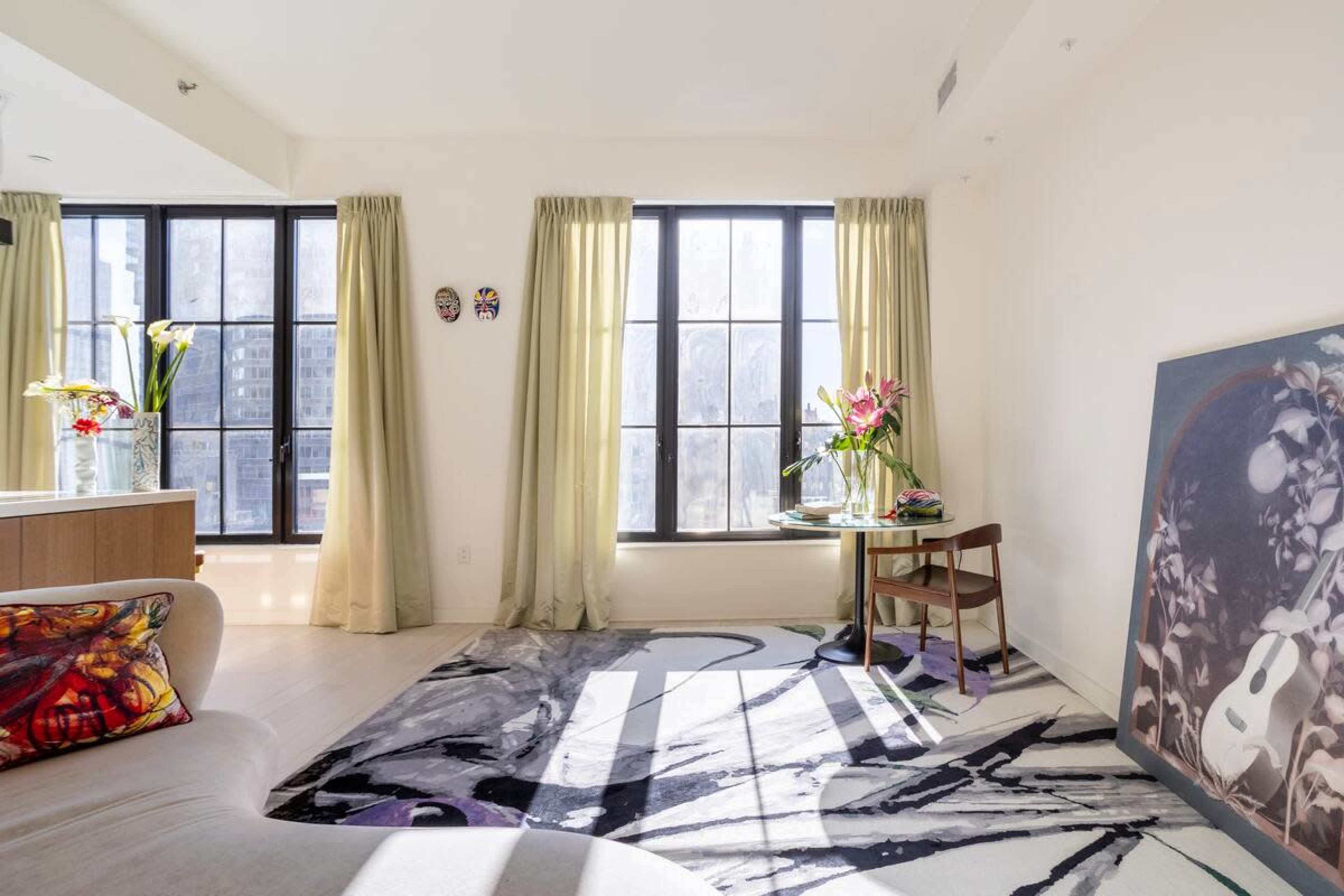 A bright living room features large windows with sheer curtains, a colorful rug, a small table with flowers, and a framed artwork leaning against the wall.