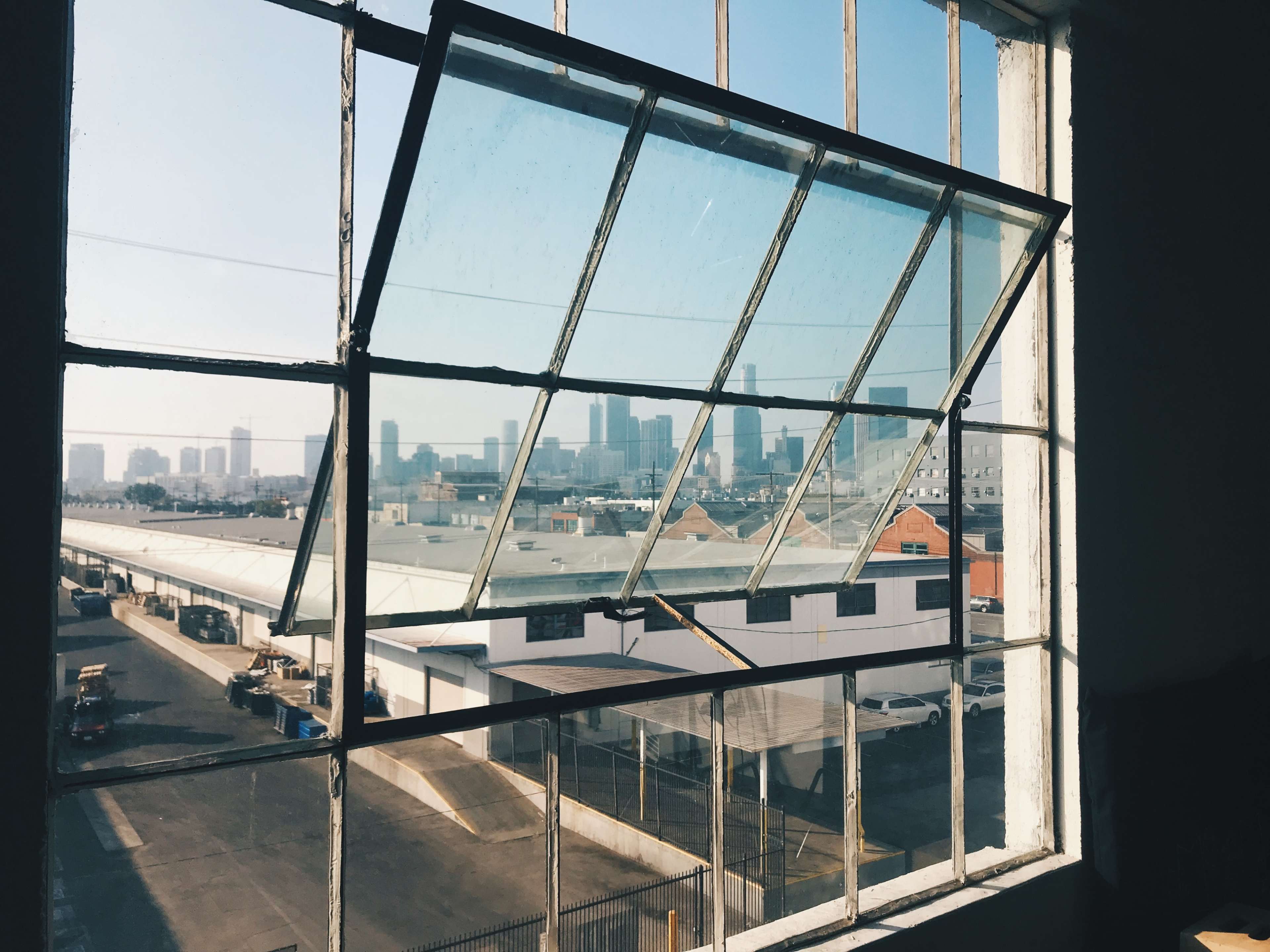 The image shows an open industrial window with multiple panes, overlooking a city skyline and an urban landscape.