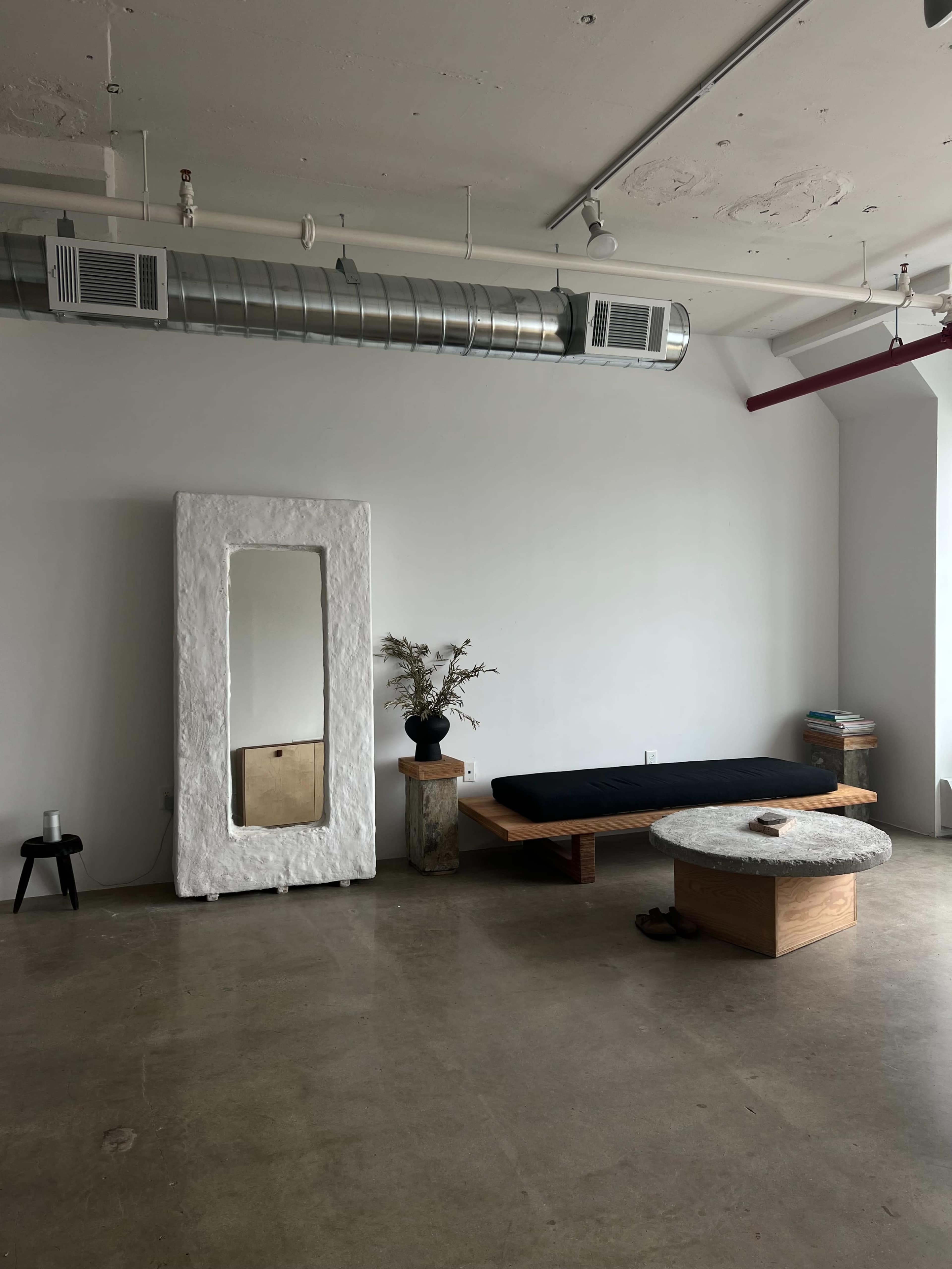 A minimalist room with a large mirror, a low wooden bed, a round coffee table, and a vase of dried flowers on a pedestal.