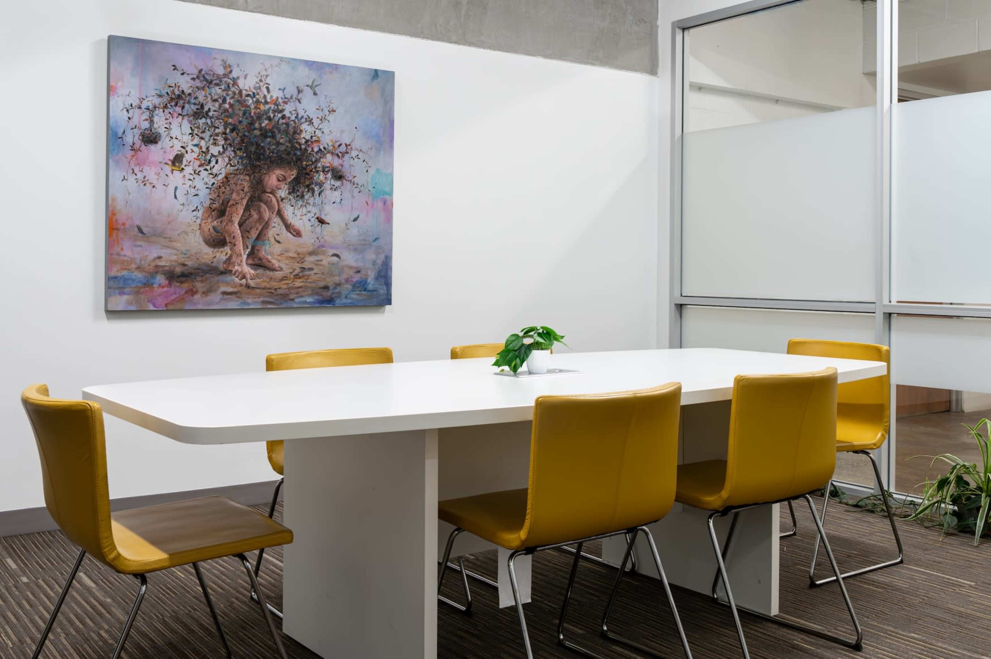 A bright meeting room features a large white table surrounded by yellow chairs, with a colorful artwork hanging on the wall.