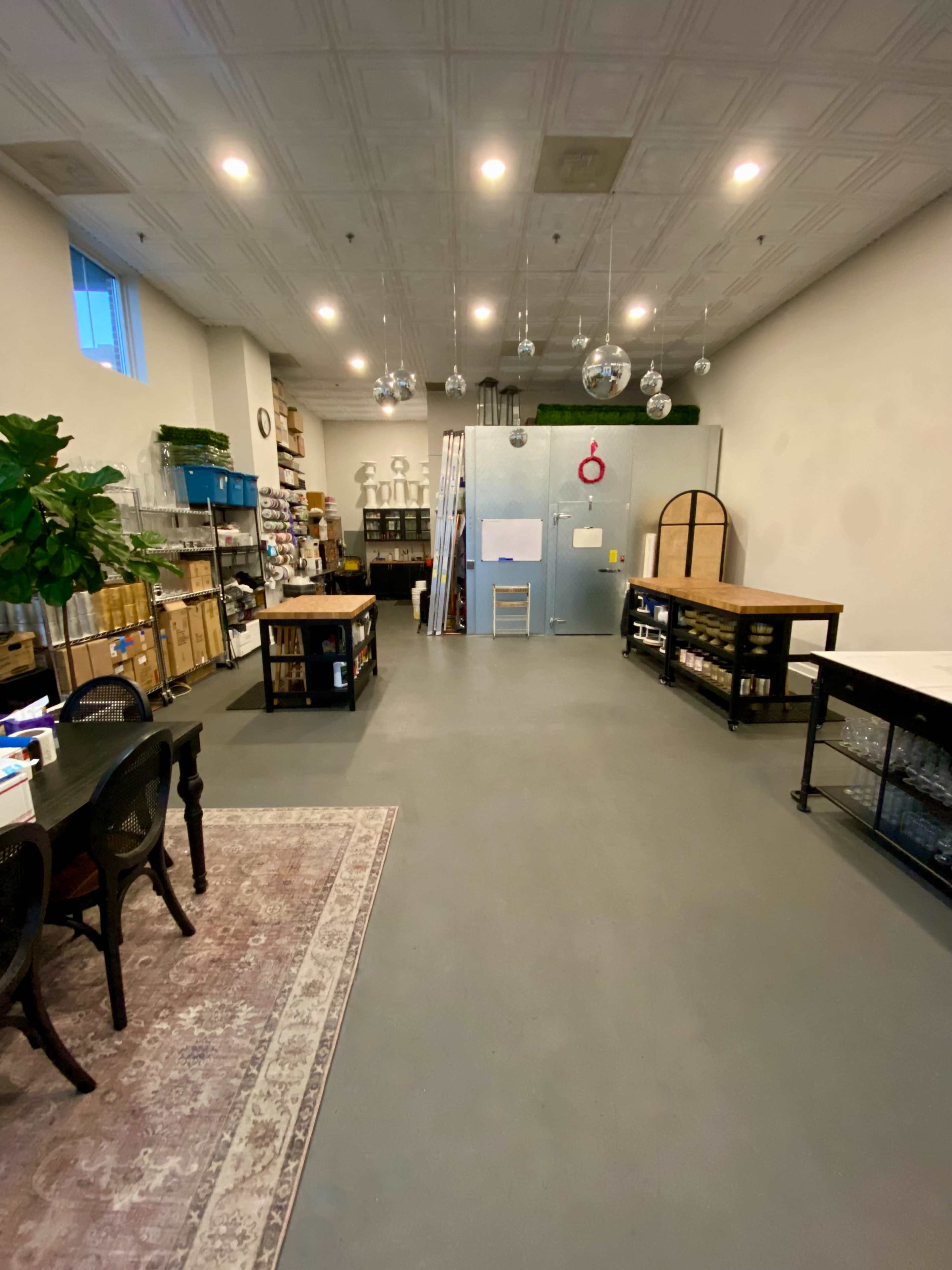 The image shows a spacious room with shelves of supplies, tables, and a large refrigerator at the back, illuminated by ceiling lights.