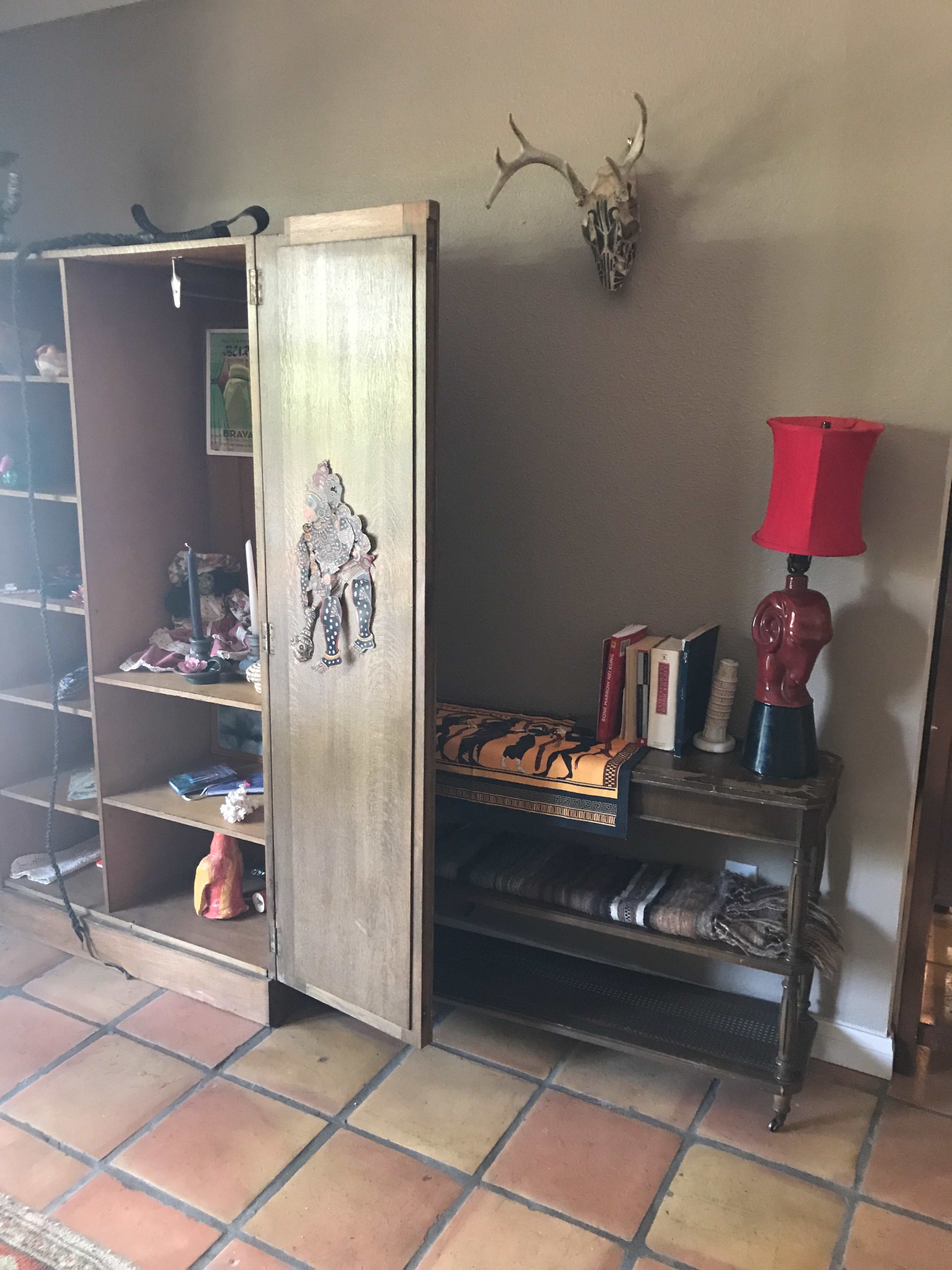 A wooden shelf unit with an open door, revealing items inside, alongside a small table with a red lamp and a collection of books.