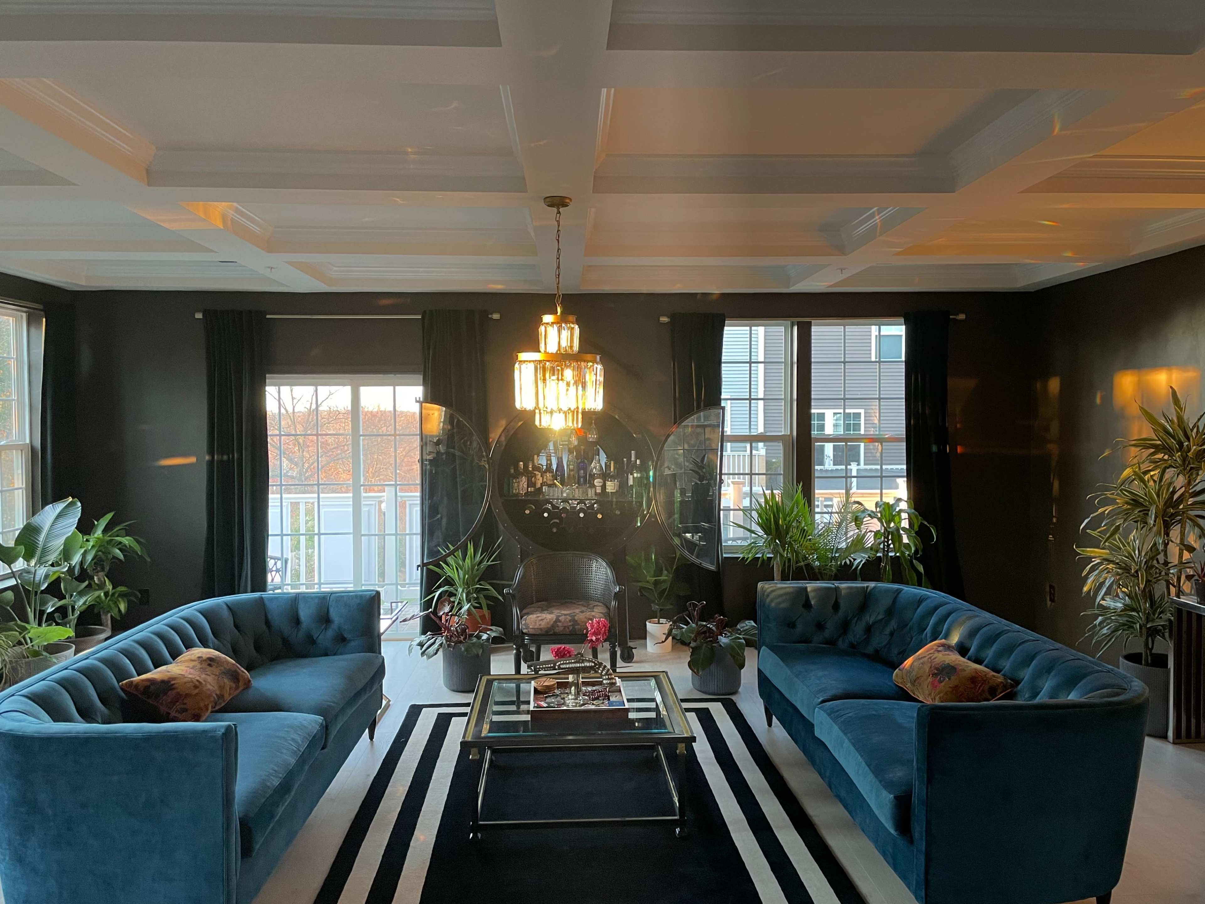 The image shows a stylish living room with two blue sofas facing each other, a coffee table in the center, and large windows surrounded by indoor plants.