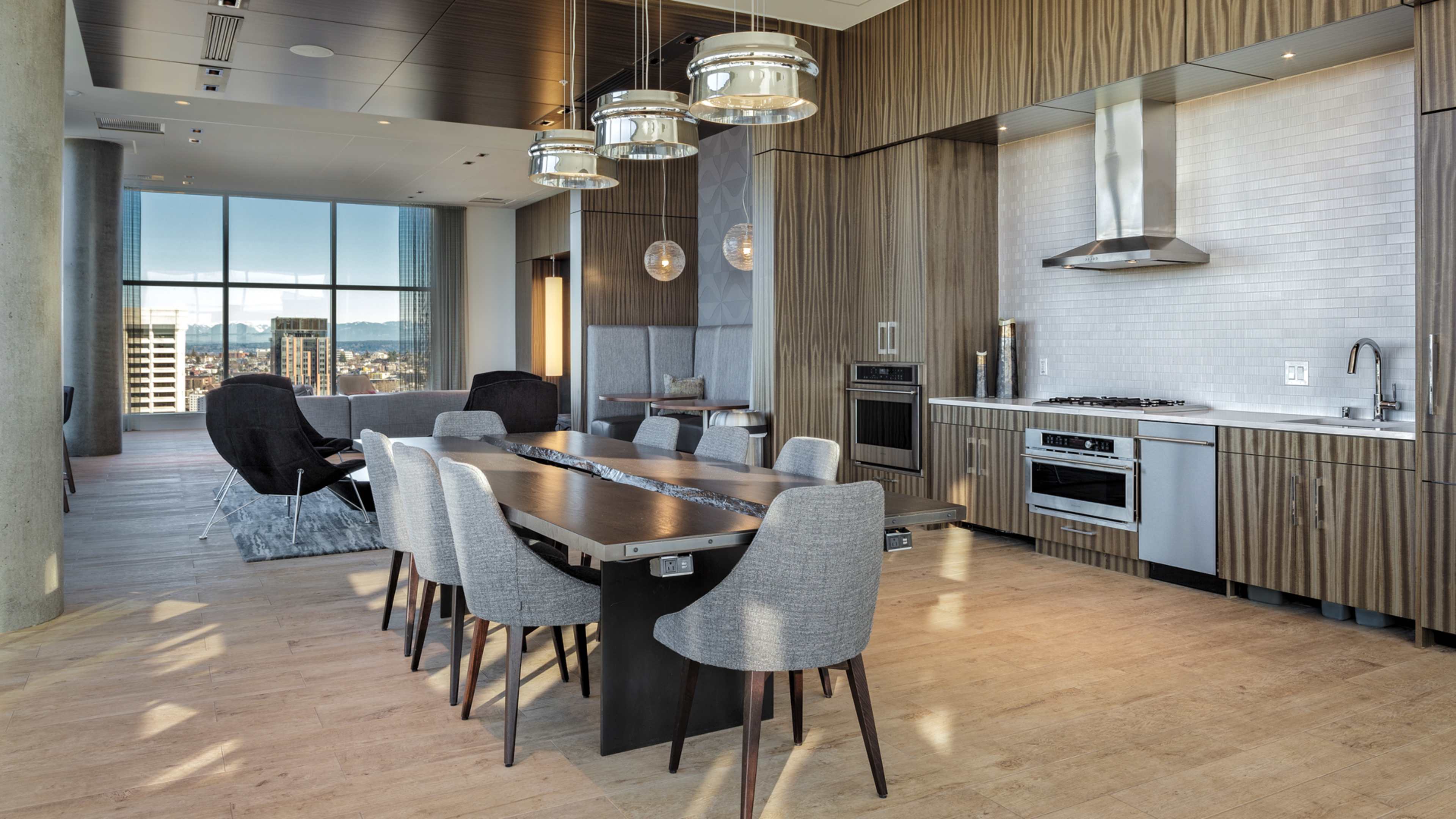 A modern dining area features a large table surrounded by gray chairs, with an adjacent kitchen equipped with stainless steel appliances and large windows offering a city view.