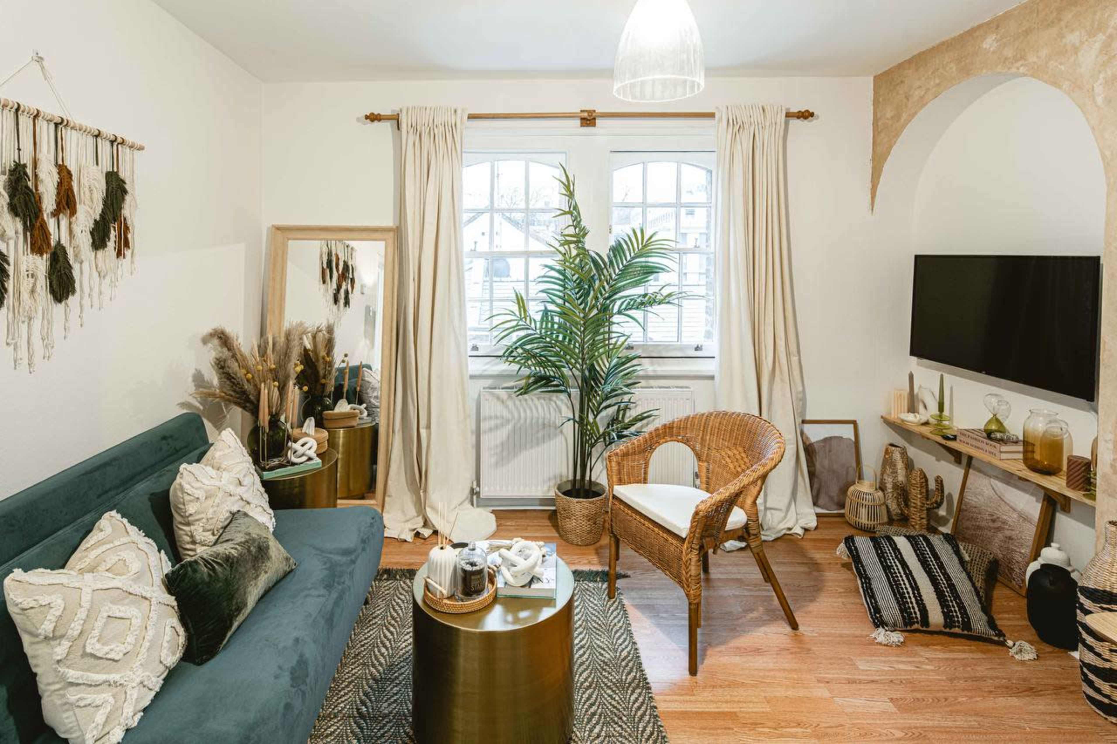 The image shows a cozy living room featuring a green sofa, a wicker chair, a large mirror, decorative plants, and a wall-mounted TV.