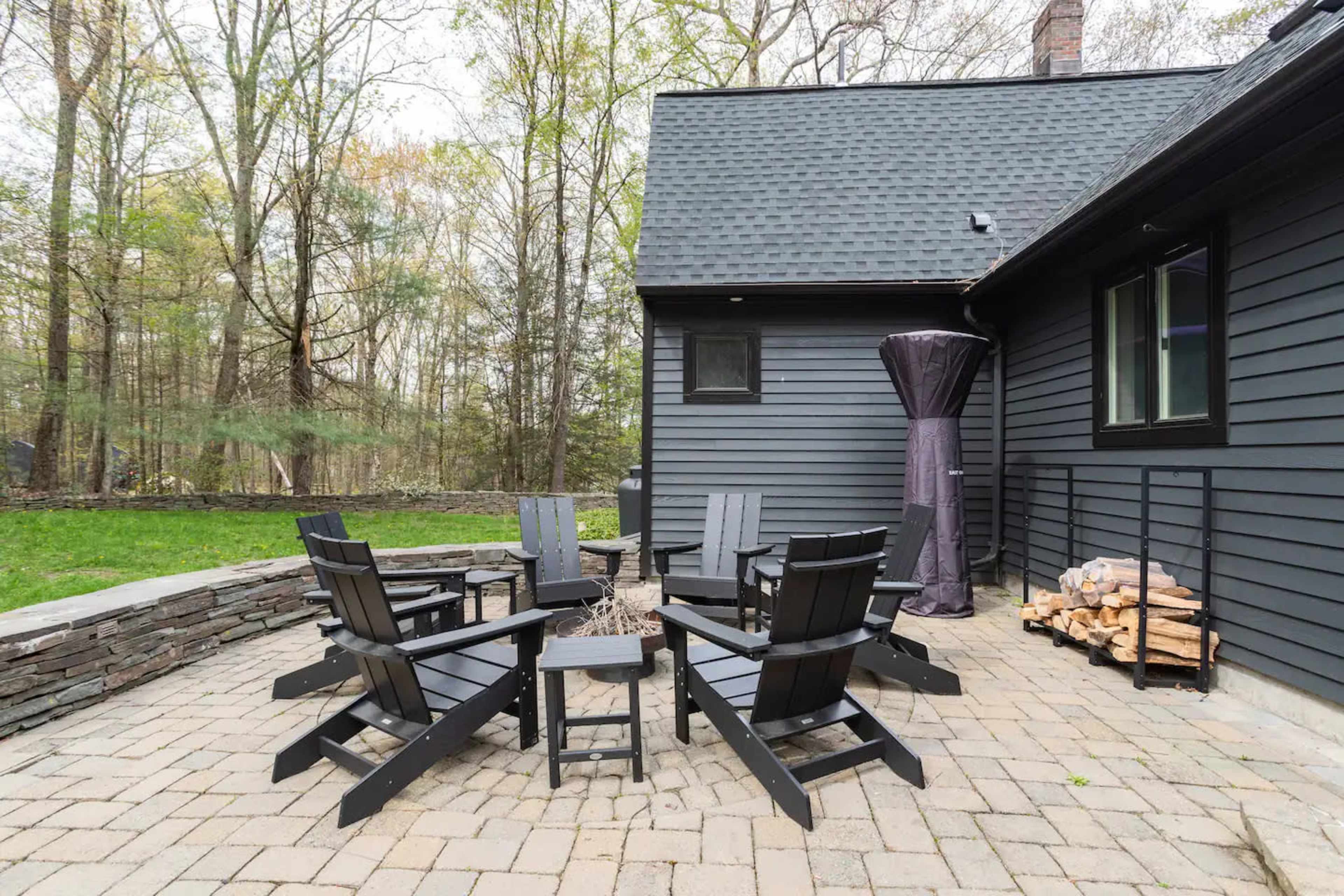 A stone patio features six black Adirondack chairs arranged around a small fire pit near a wooded area.