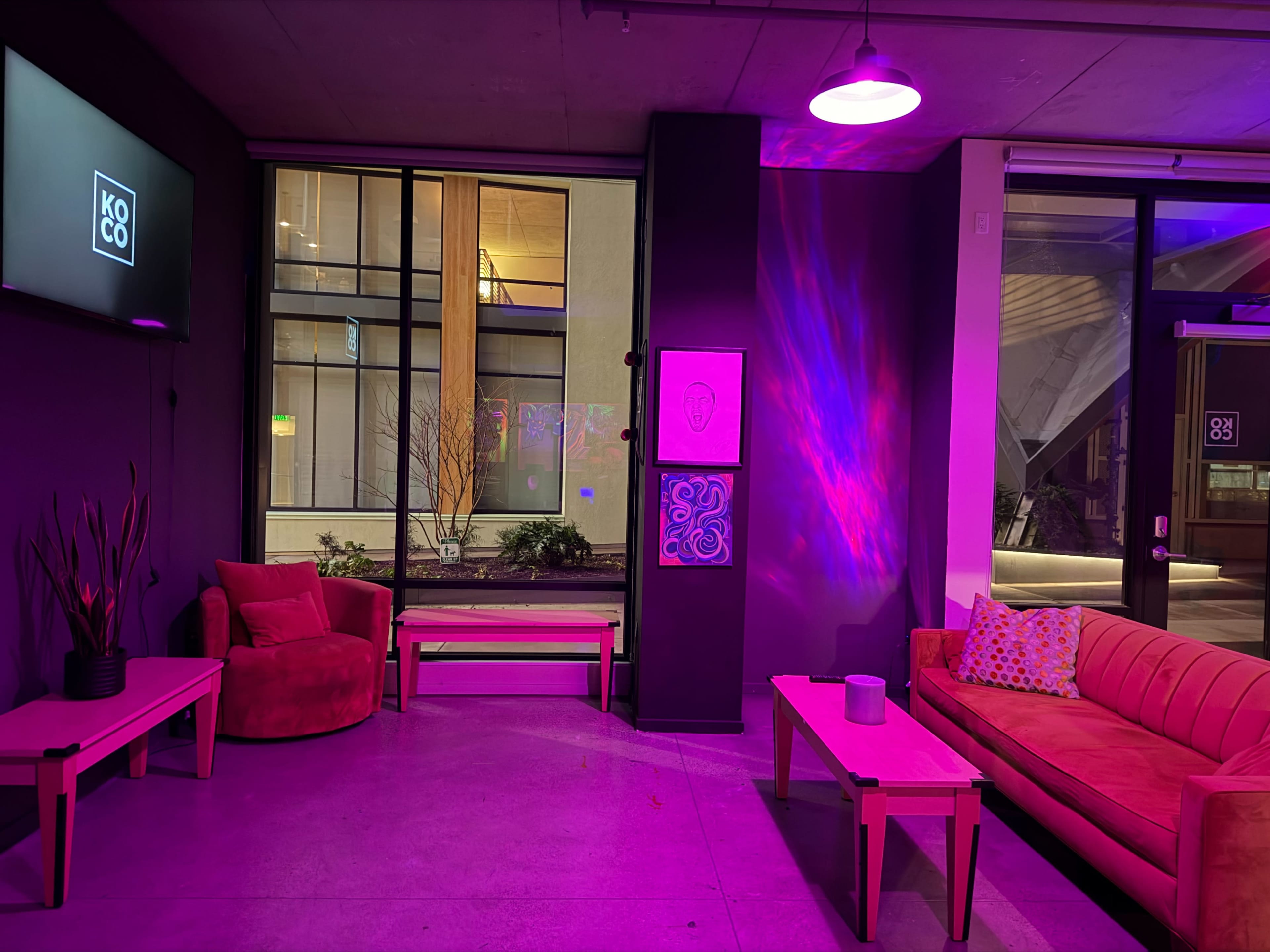 The image shows a colorful lounge area with pink lighting, featuring a red couch, two chairs, and artwork on the walls.