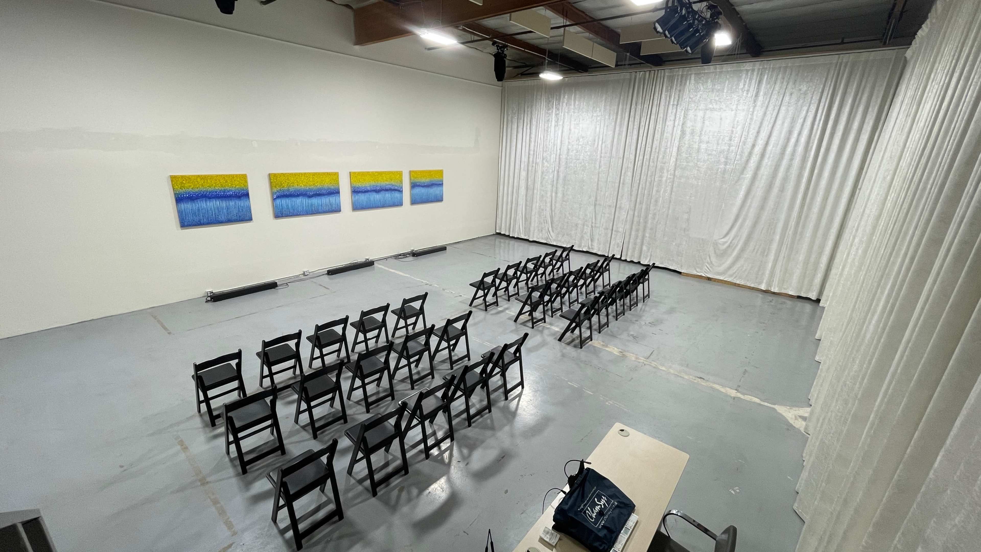 An empty room set up for an event, featuring rows of black chairs facing a set of three colorful paintings on the wall, with white curtains draped across one side.