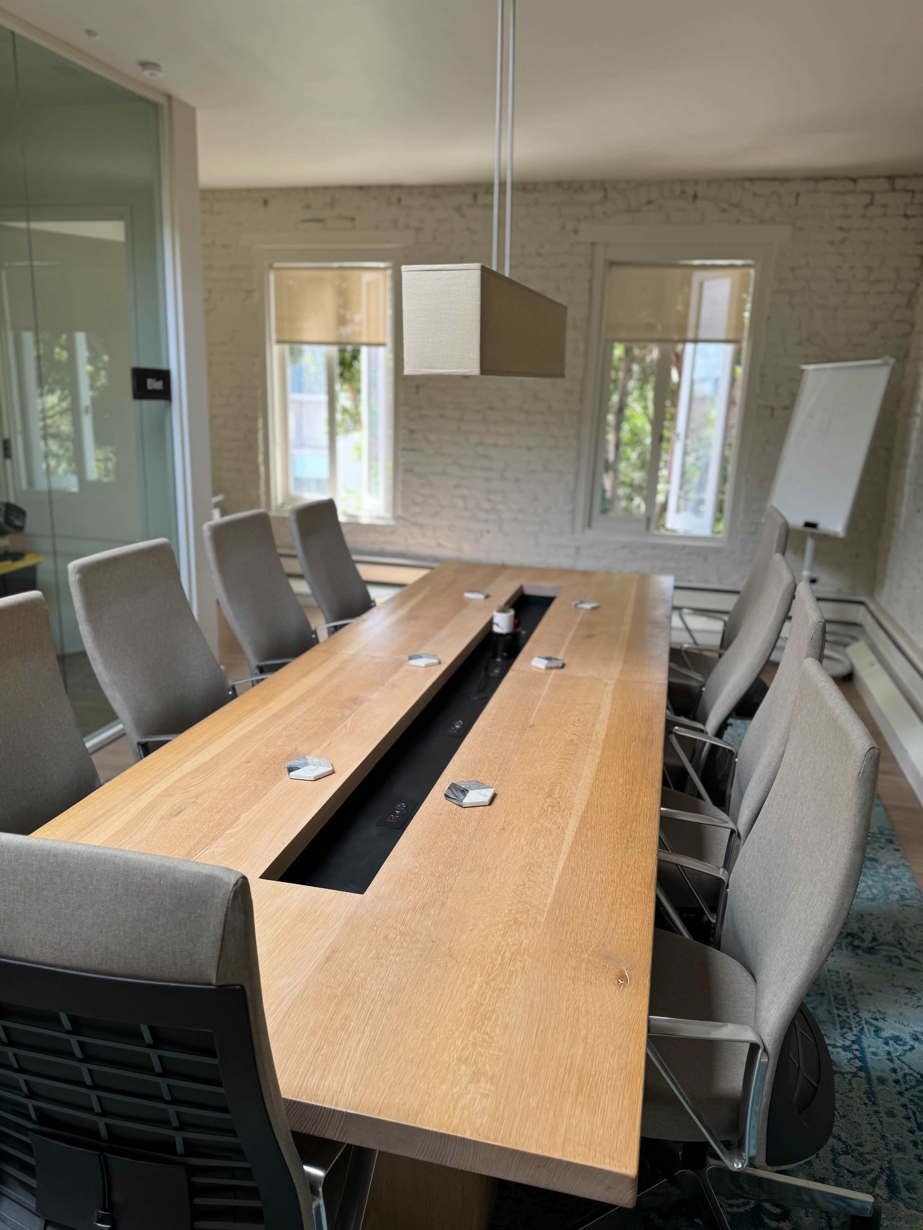A large wooden conference table with eight chairs is positioned in a bright meeting room featuring three windows and a presentation board.