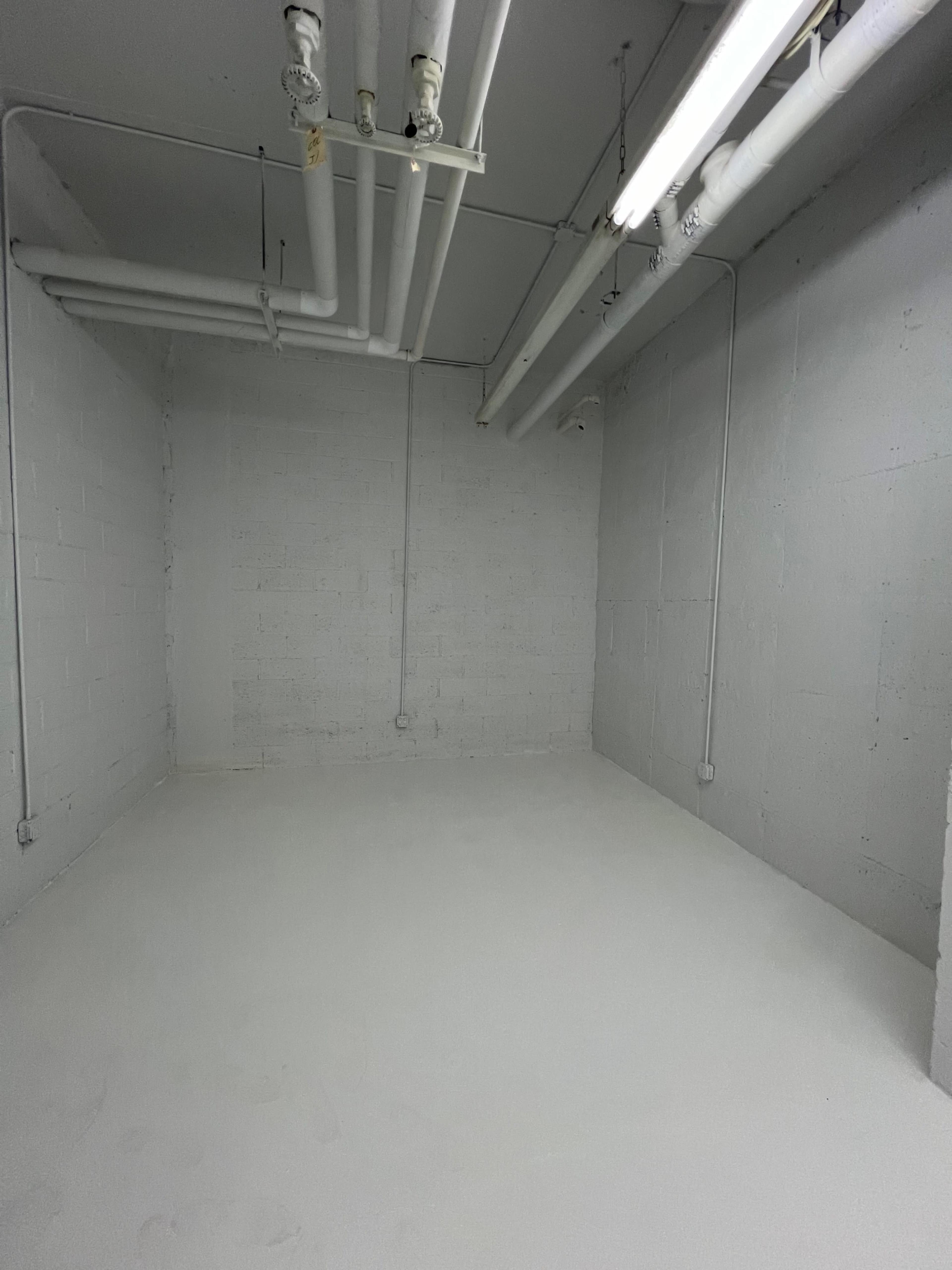A small, empty room with gray concrete walls and a white floor, illuminated by overhead fluorescent lights.