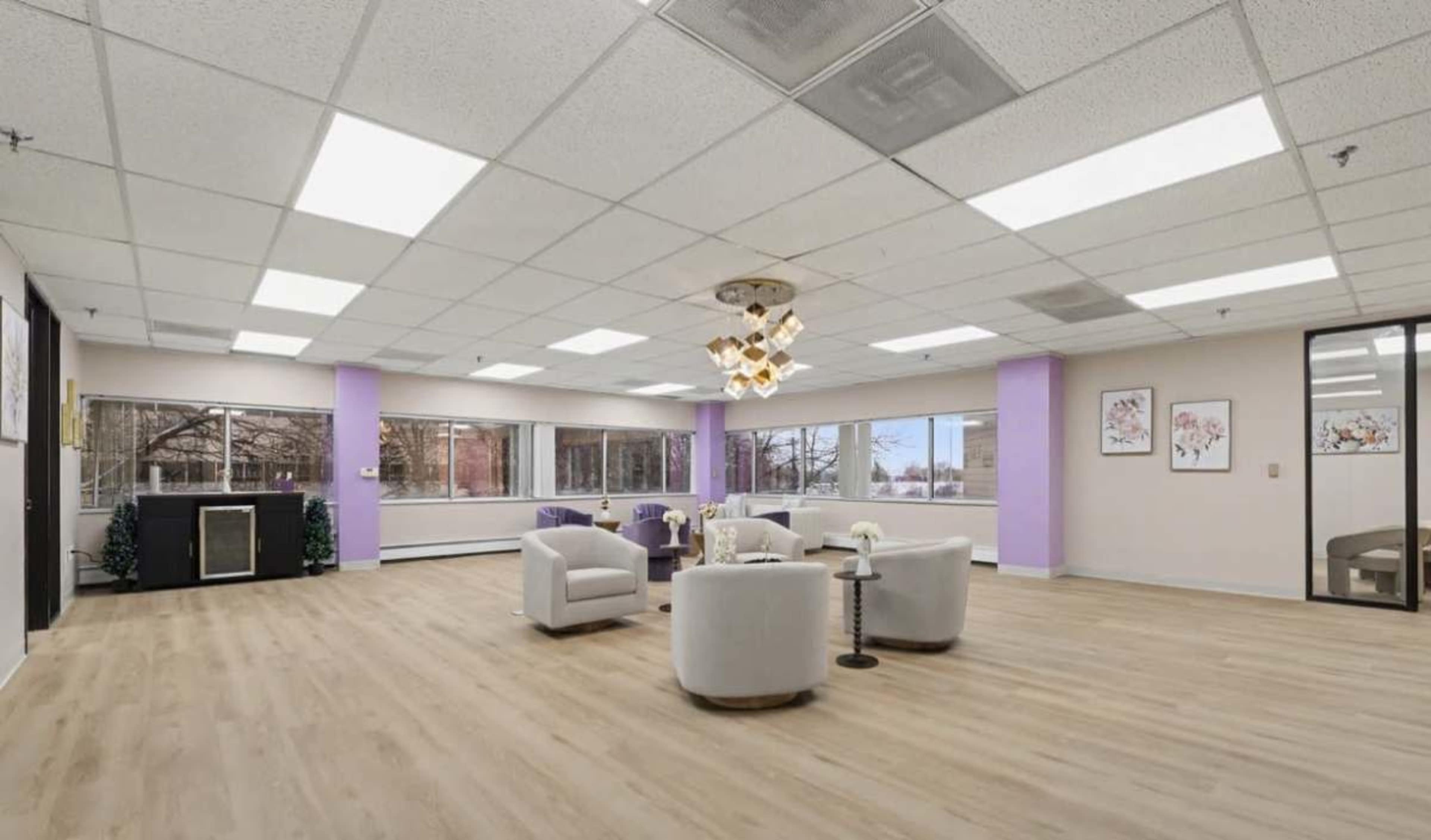 The image shows a spacious, well-lit room with light-colored flooring and purple accent walls, furnished with white armchairs, a coffee table, and decorated with large windows.