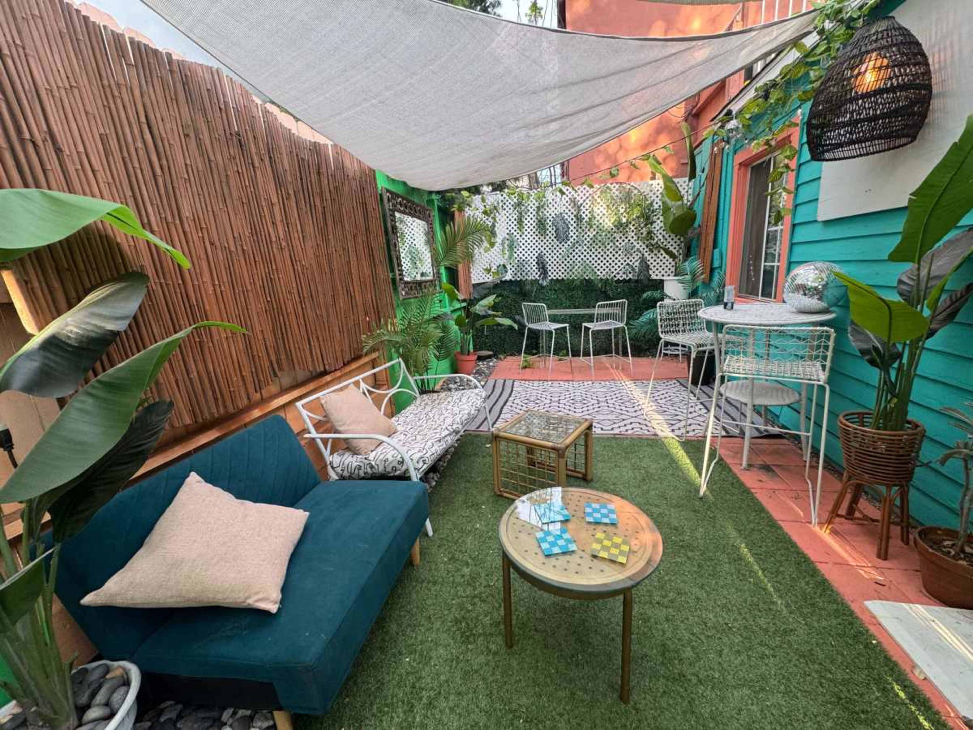 The image shows a cozy outdoor patio with green grass, various plants, a blue sofa, a coffee table, and a shaded seating area.