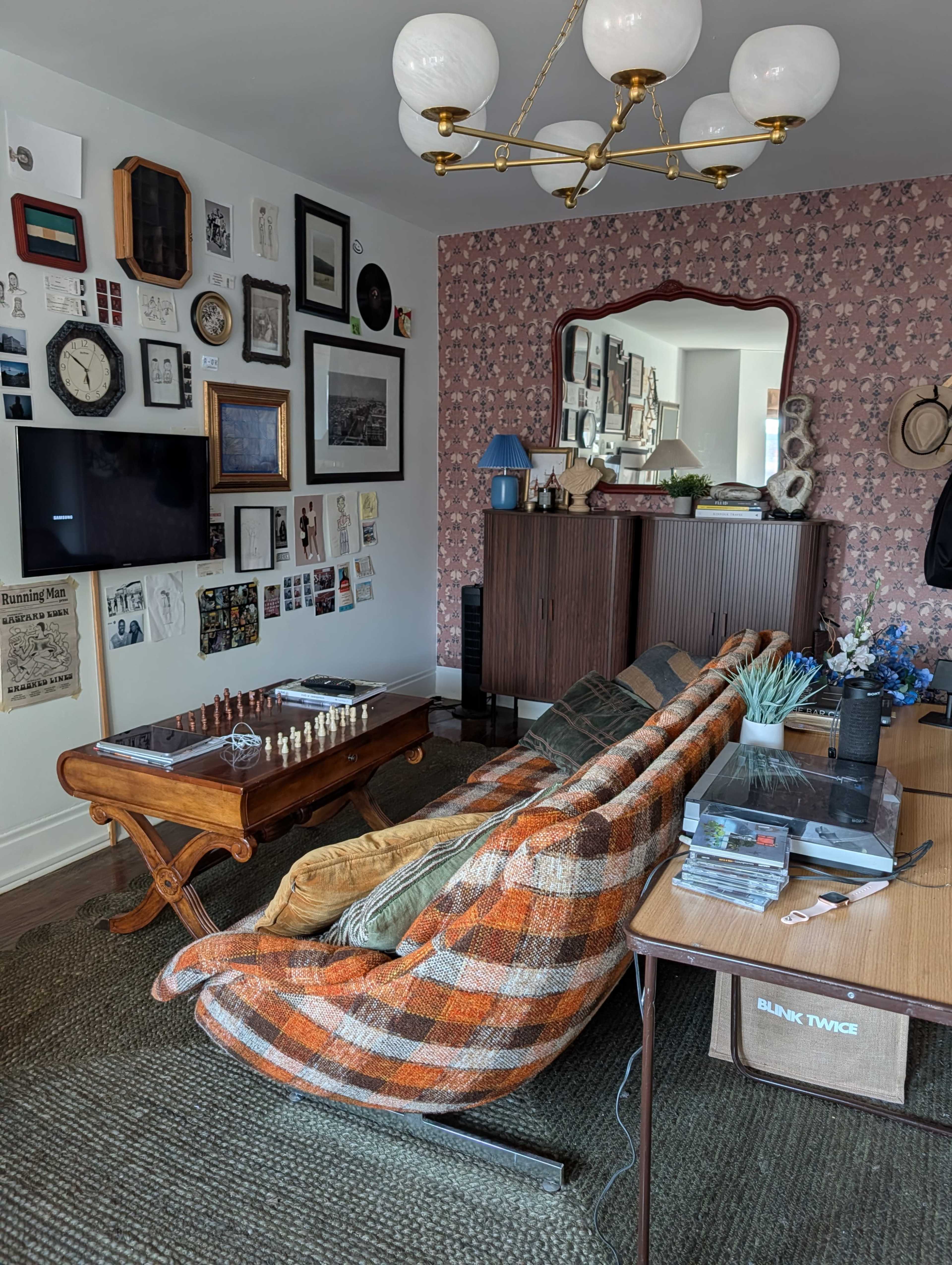 The image shows a living room with an eclectic mix of wall art, a checkered sofa, a wooden coffee table with a chess set, and a vintage-style cabinet under a chandelier.