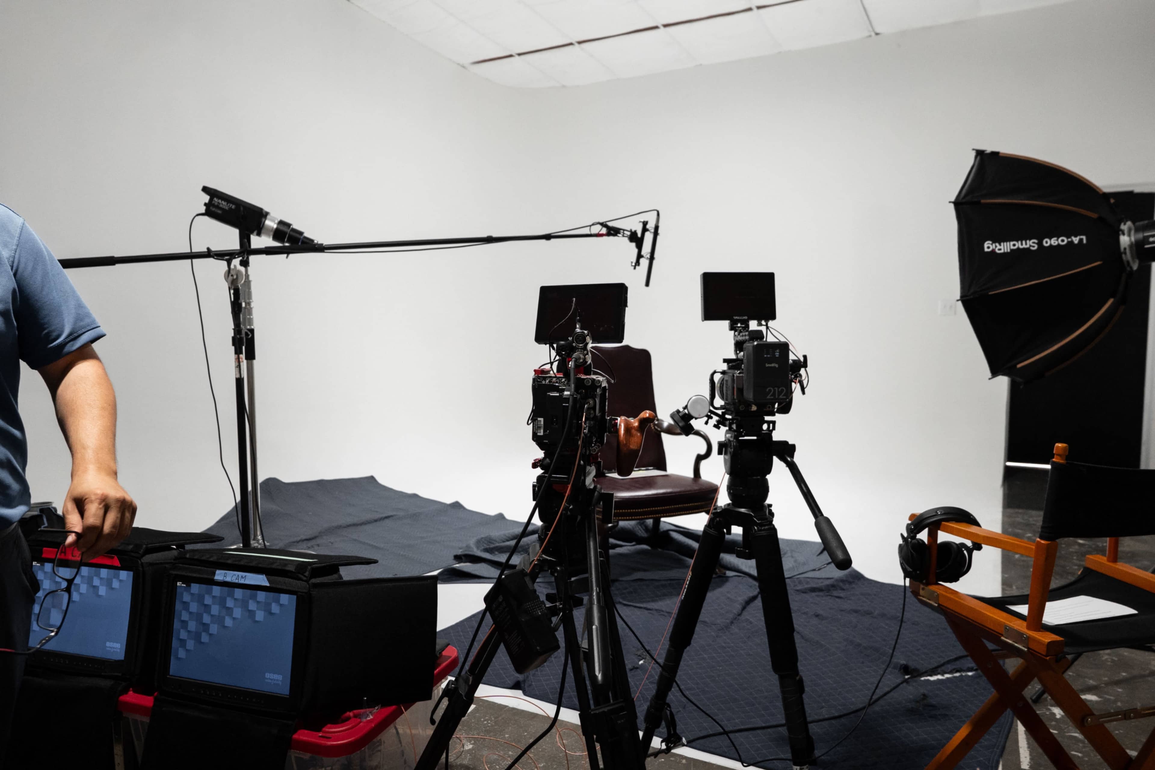 The image shows a video production setup featuring multiple cameras, microphones, and a studio chair placed on a black mat in front of a plain white backdrop.