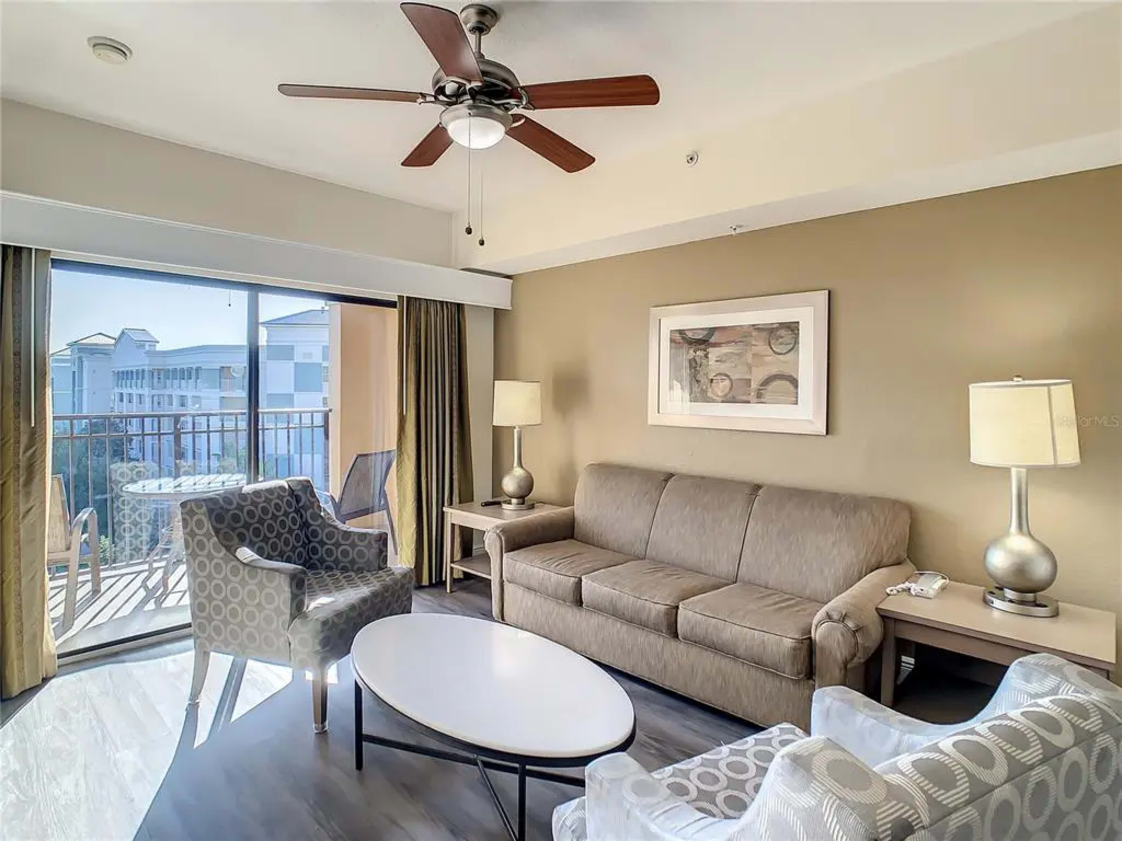 A living room with a sofa, two patterned chairs, a round coffee table, a ceiling fan, and a balcony door leading to an outdoor view.