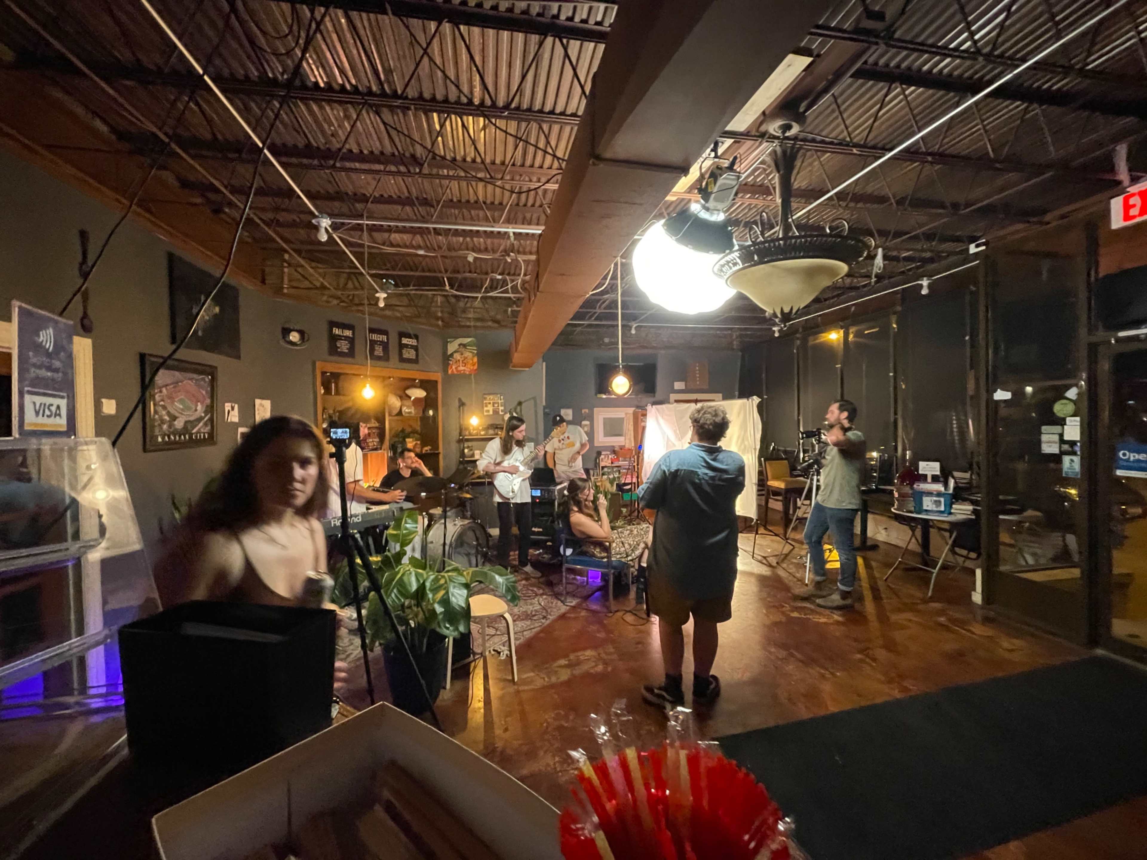 A dimly lit café with several people seated and engaged in conversation, while one person stands near the entrance.